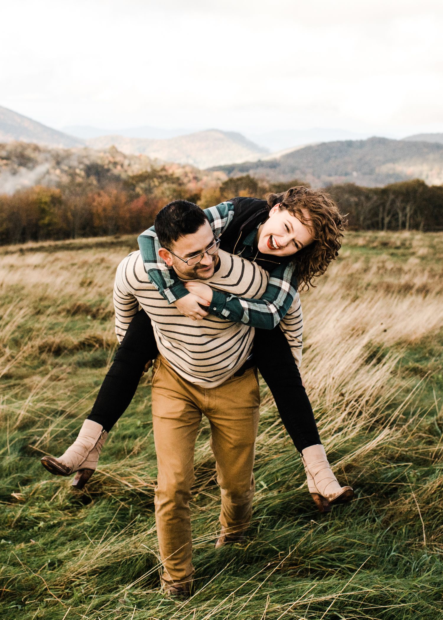 Laughing woman getting a piggyback ride from man in striped sweater in an open mountain meadow.
