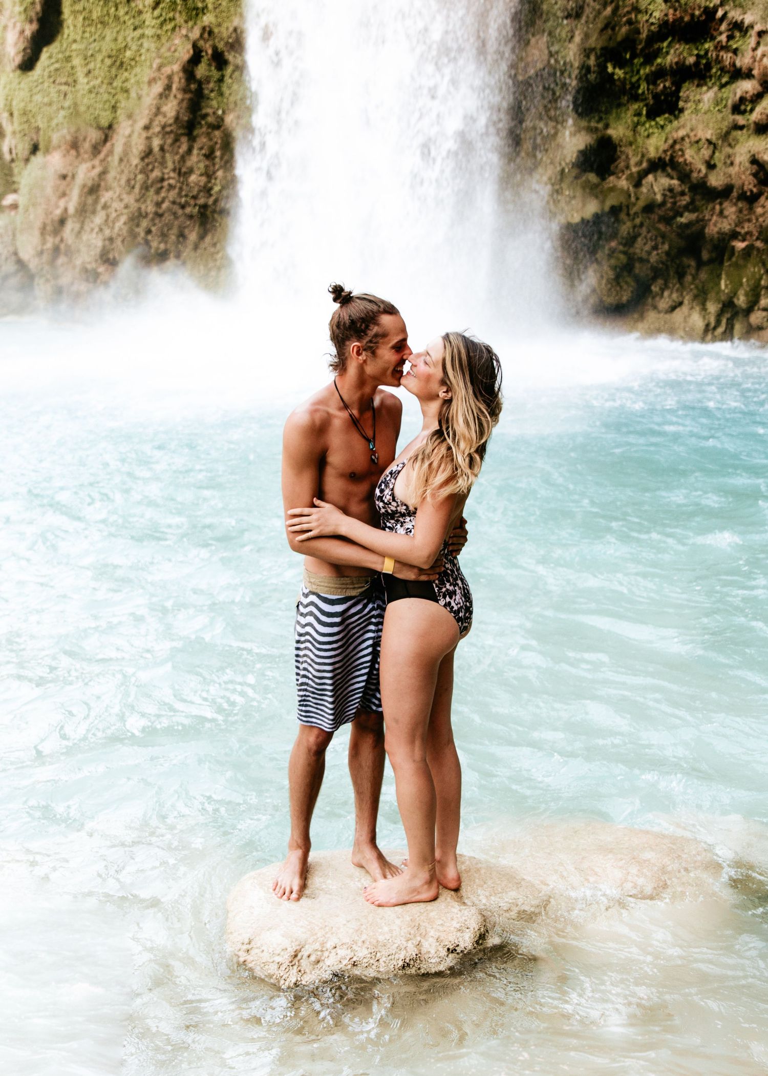 Couple kissing on a rock in turquoise water with a majestic waterfall behind them.