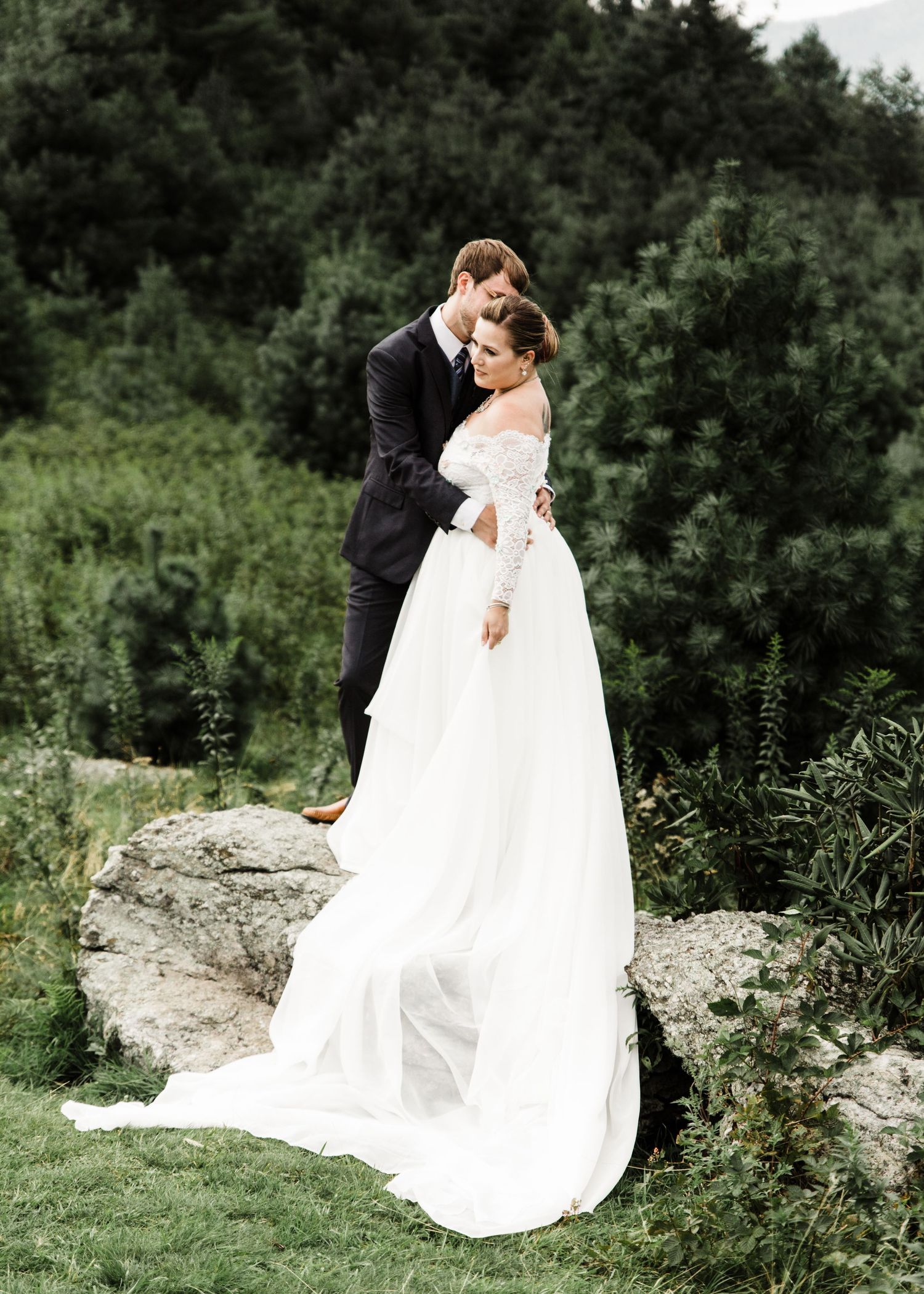 Bride in off-shoulder lace gown and groom in dark suit posing on rocks with evergreen trees behind.