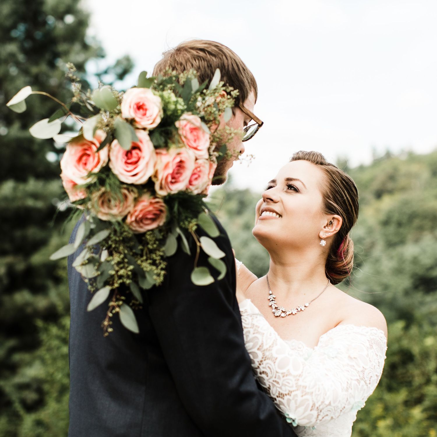 Smiling bride in lace gown embraces groom holding large pink rose and greenery bouquet outdoors.