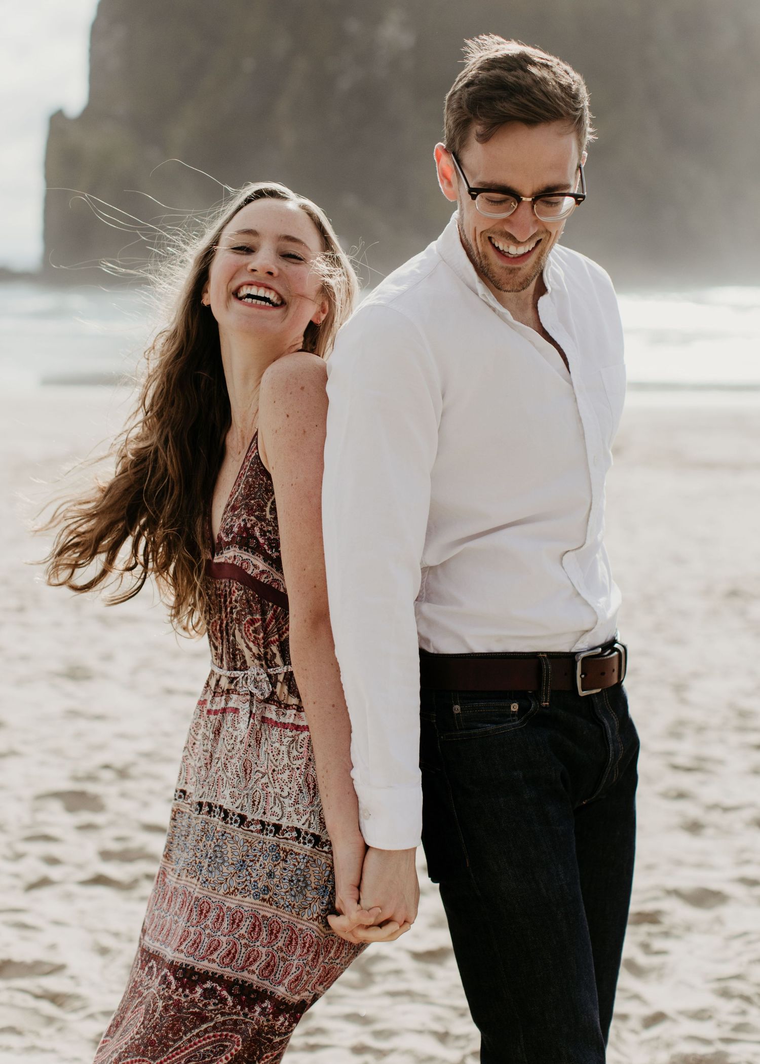 Couple laughing and holding hands on a beach with dramatic rock formations in the background.