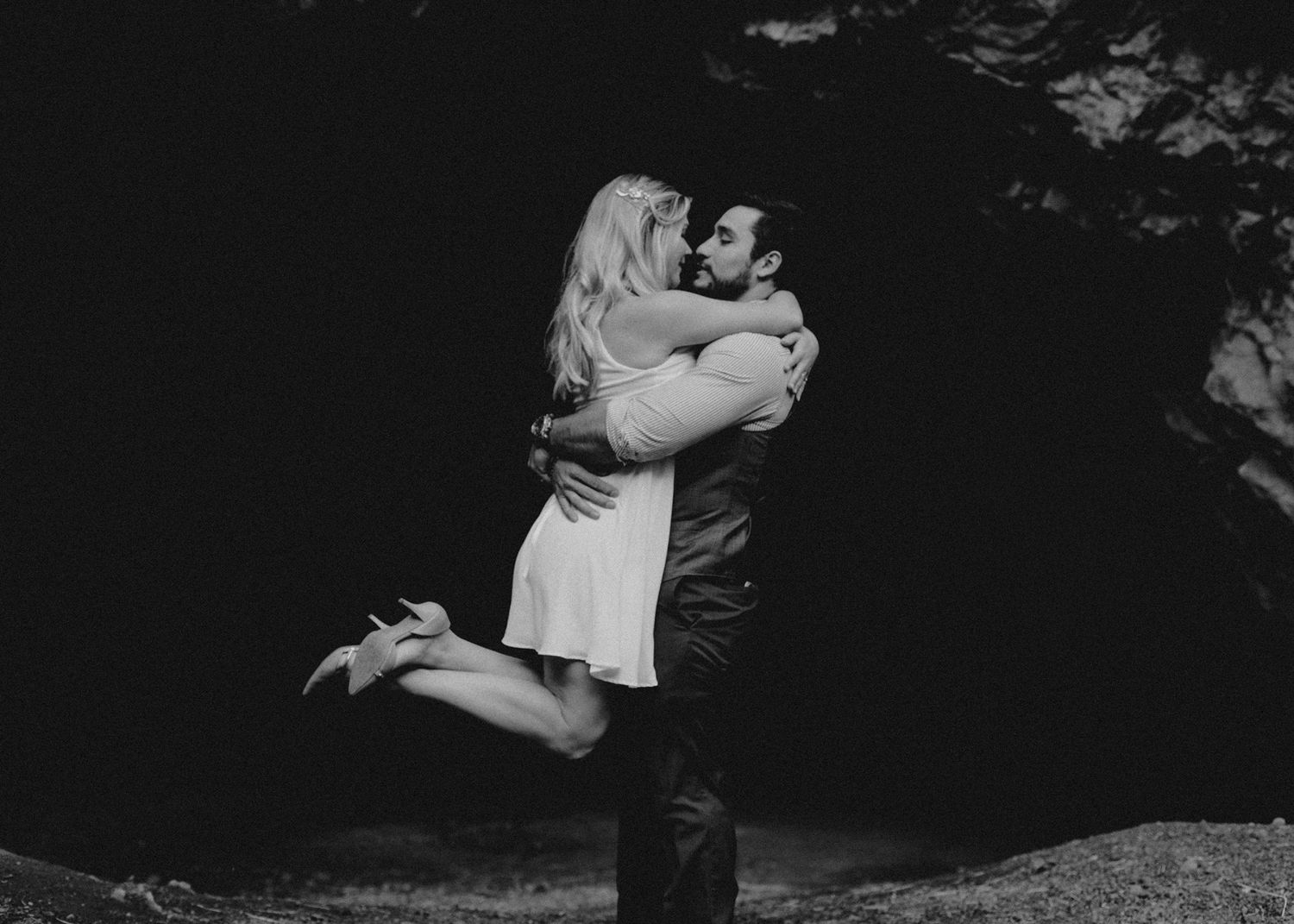 Black and white photo of couple embracing romantically in front of a dark rocky backdrop outdoors.