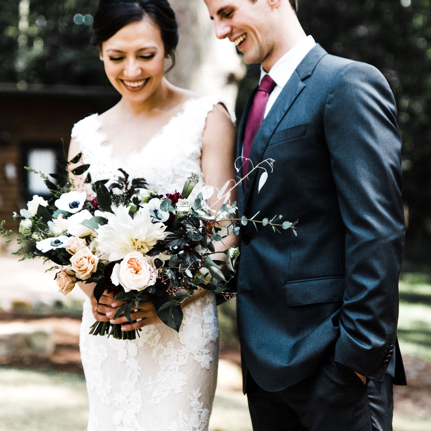 Bride in lace gown holding floral bouquet smiling with groom in gray suit.