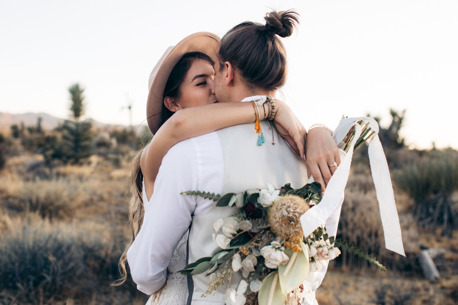 Two brides embracing in desert landscape, one holding a boho bouquet.