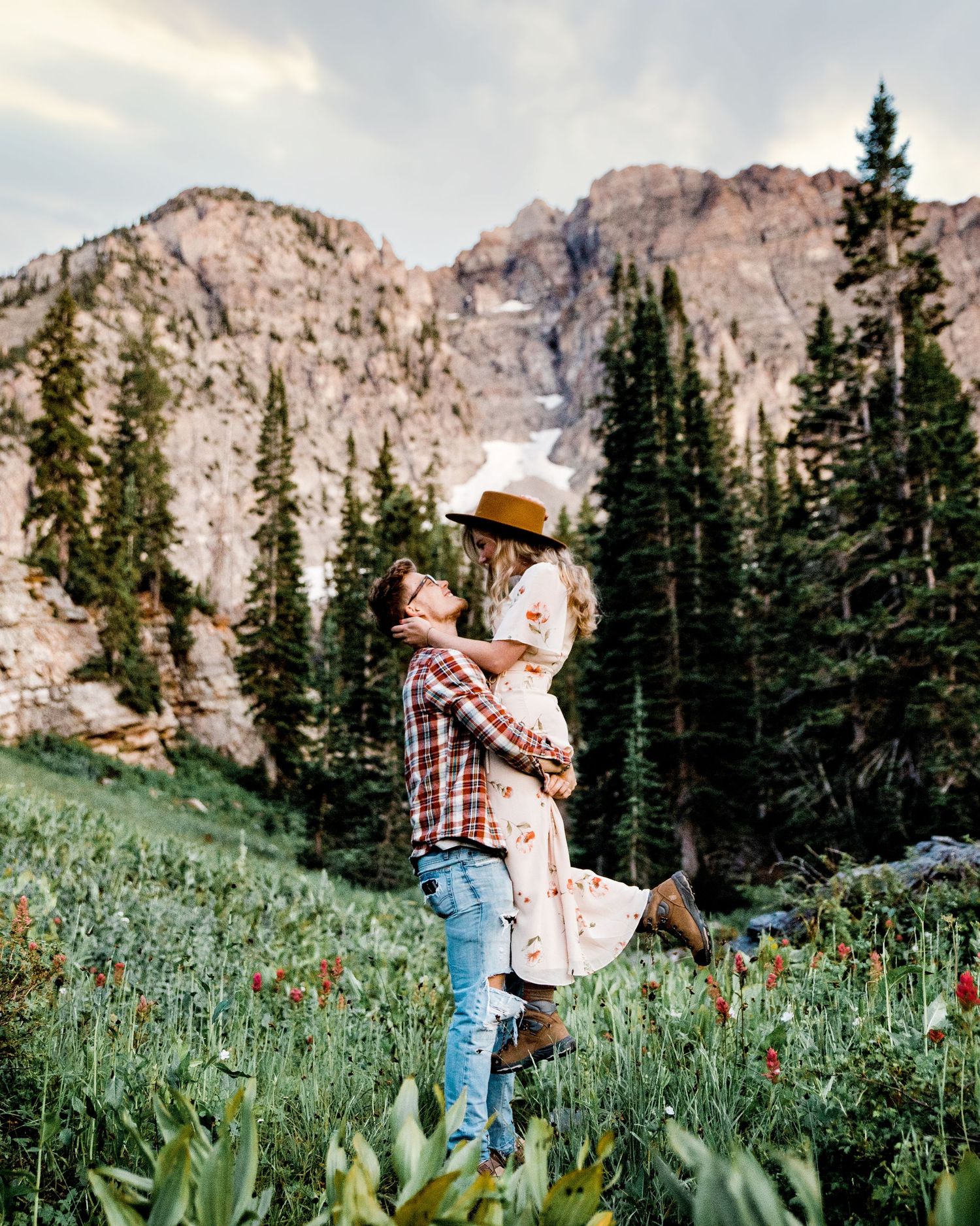 Man lifting woman in a floral dress in a colorful mountain wildflower meadow at dusk.