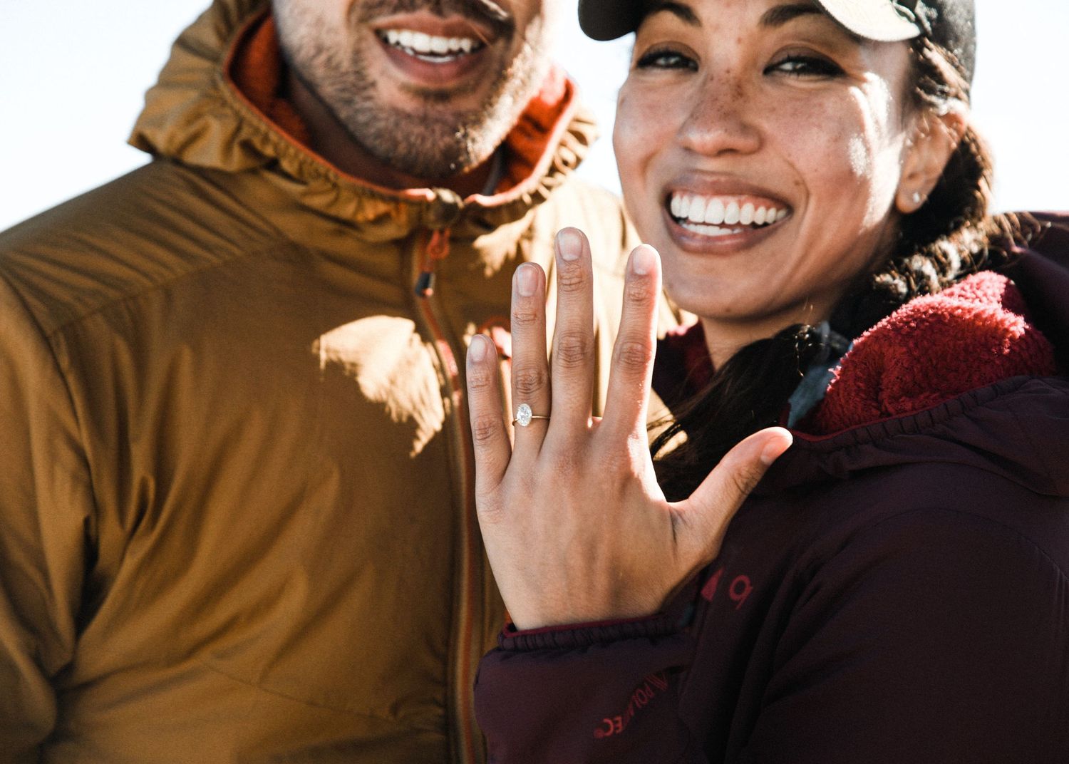 Smiling couple displaying diamond engagement ring with snowy mountain backdrop in winter gear.