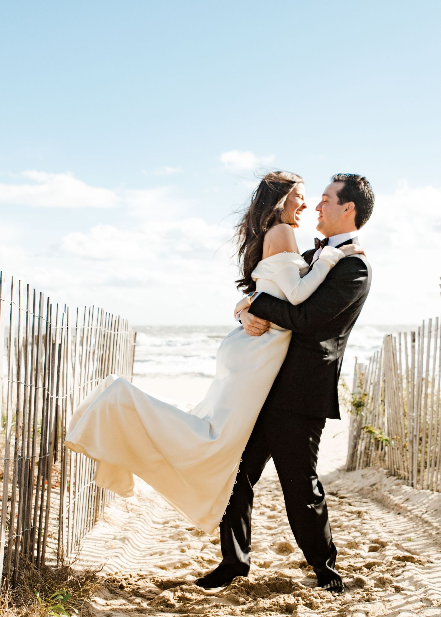 Groom joyfully lifting his laughing bride in a white off-shoulder gown on a sunny beach wedding day.