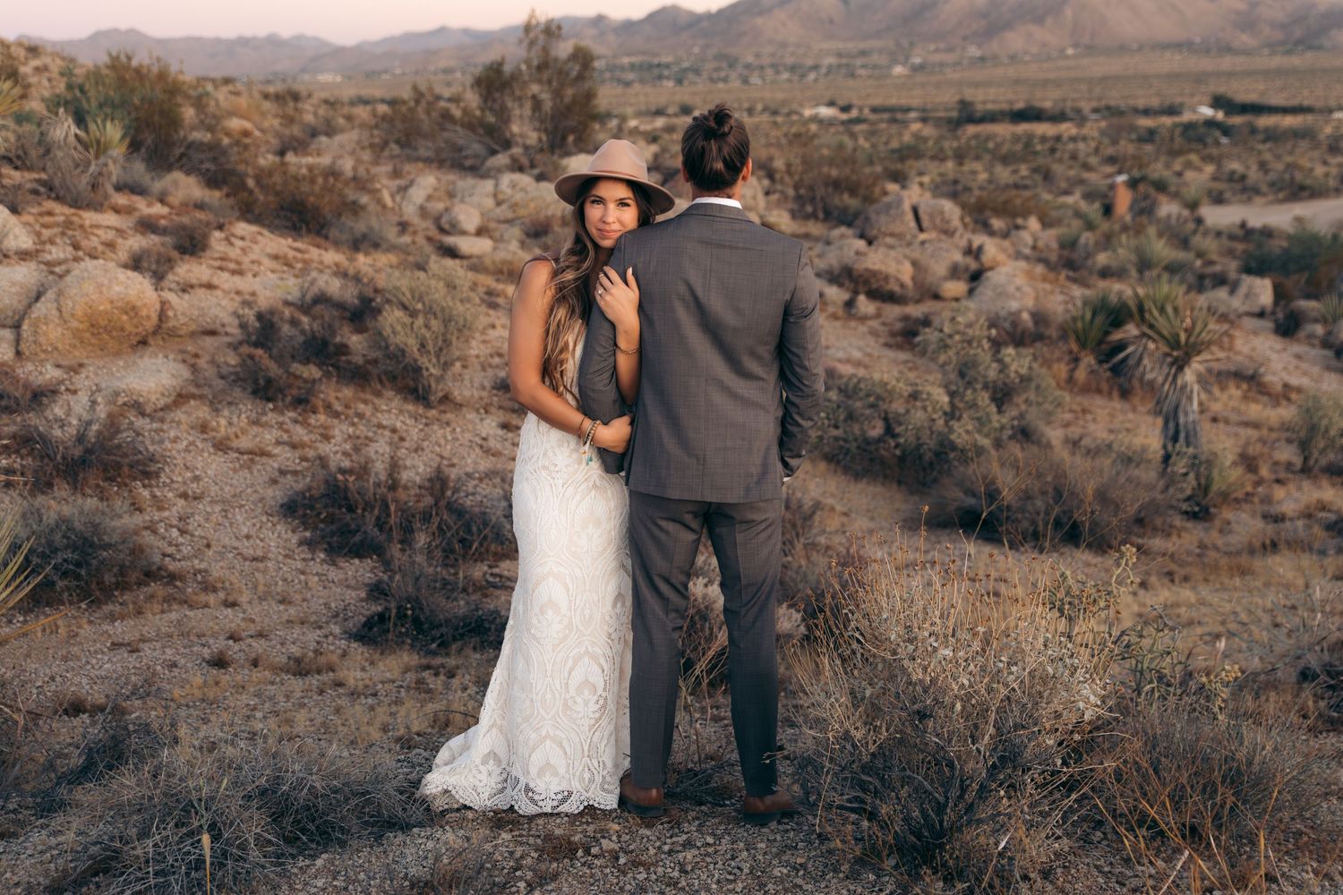A bride in a lace wedding dress and hat stands with her groom in a scenic desert landscape at sunset.