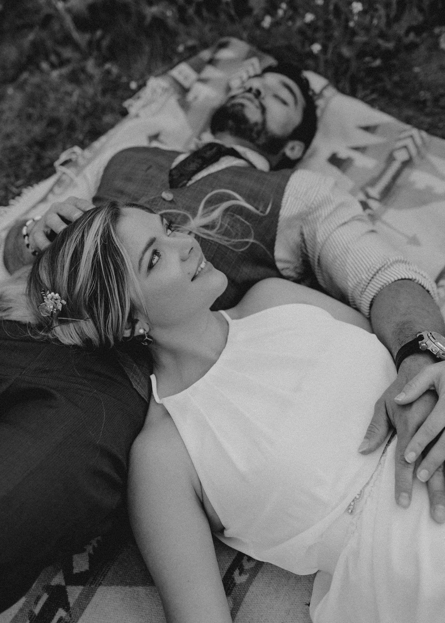 Black and white photo of a couple lying on a blanket outdoors, holding hands in a romantic moment.