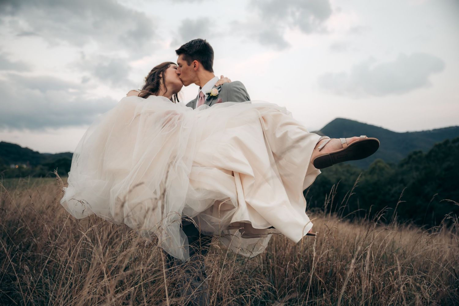Groom lifting bride in flowing gown in golden field with mountain views.