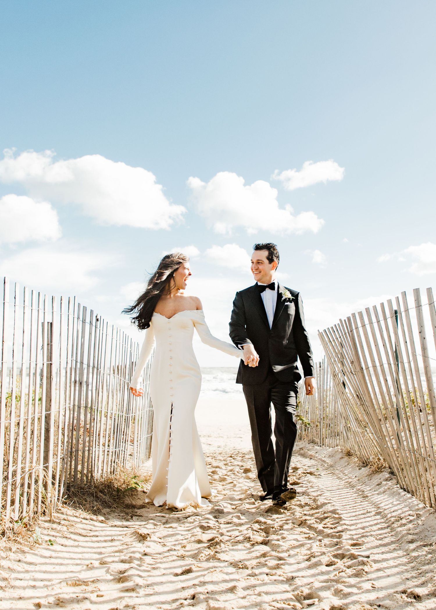 Elegant bride and groom walk hand-in-hand on a sunny sandy beach path flanked by wooden fences.
