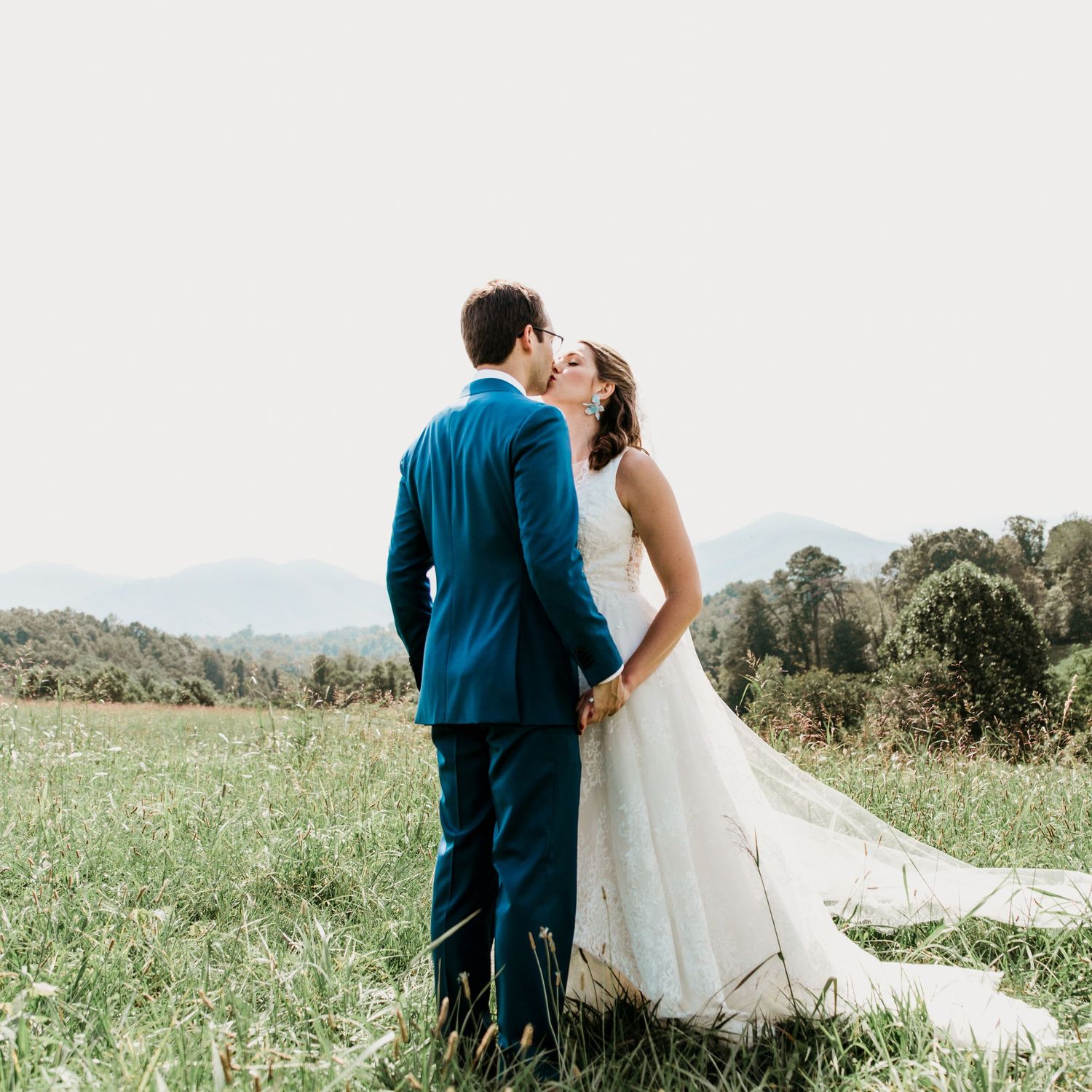 Bride and groom share a kiss while holding hands in a lush green field with mountain views behind them.