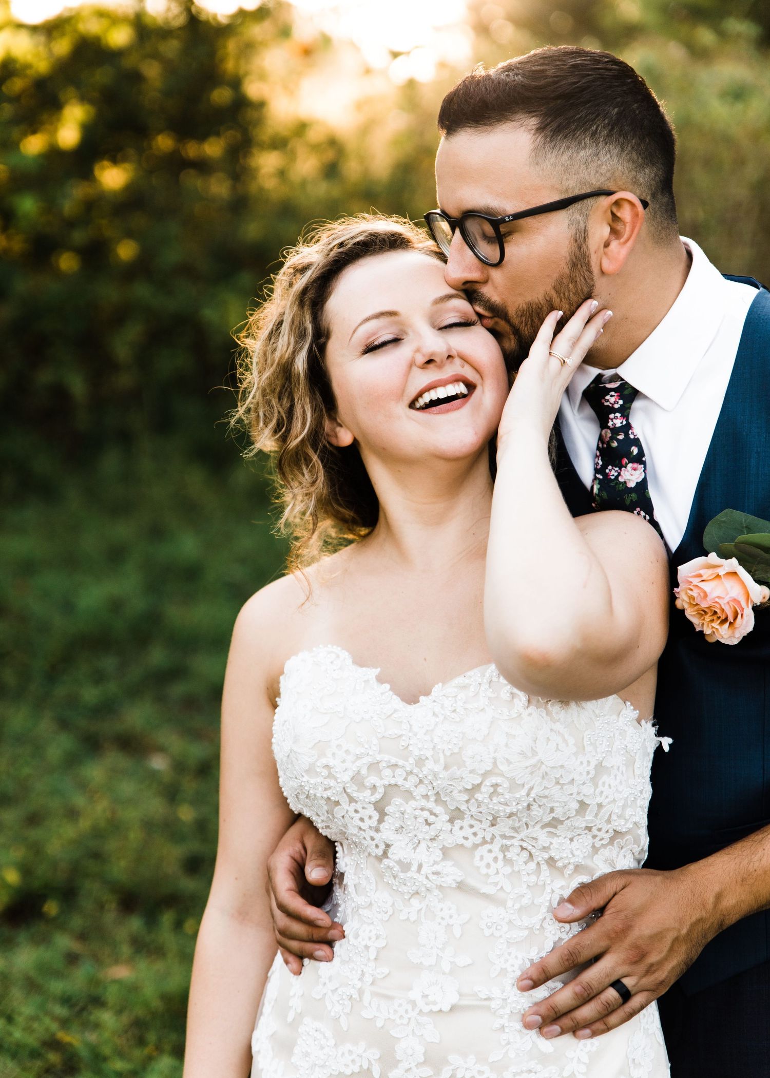 Joyful bride in lace gown laughs as groom in navy suit kisses her cheek outdoors.