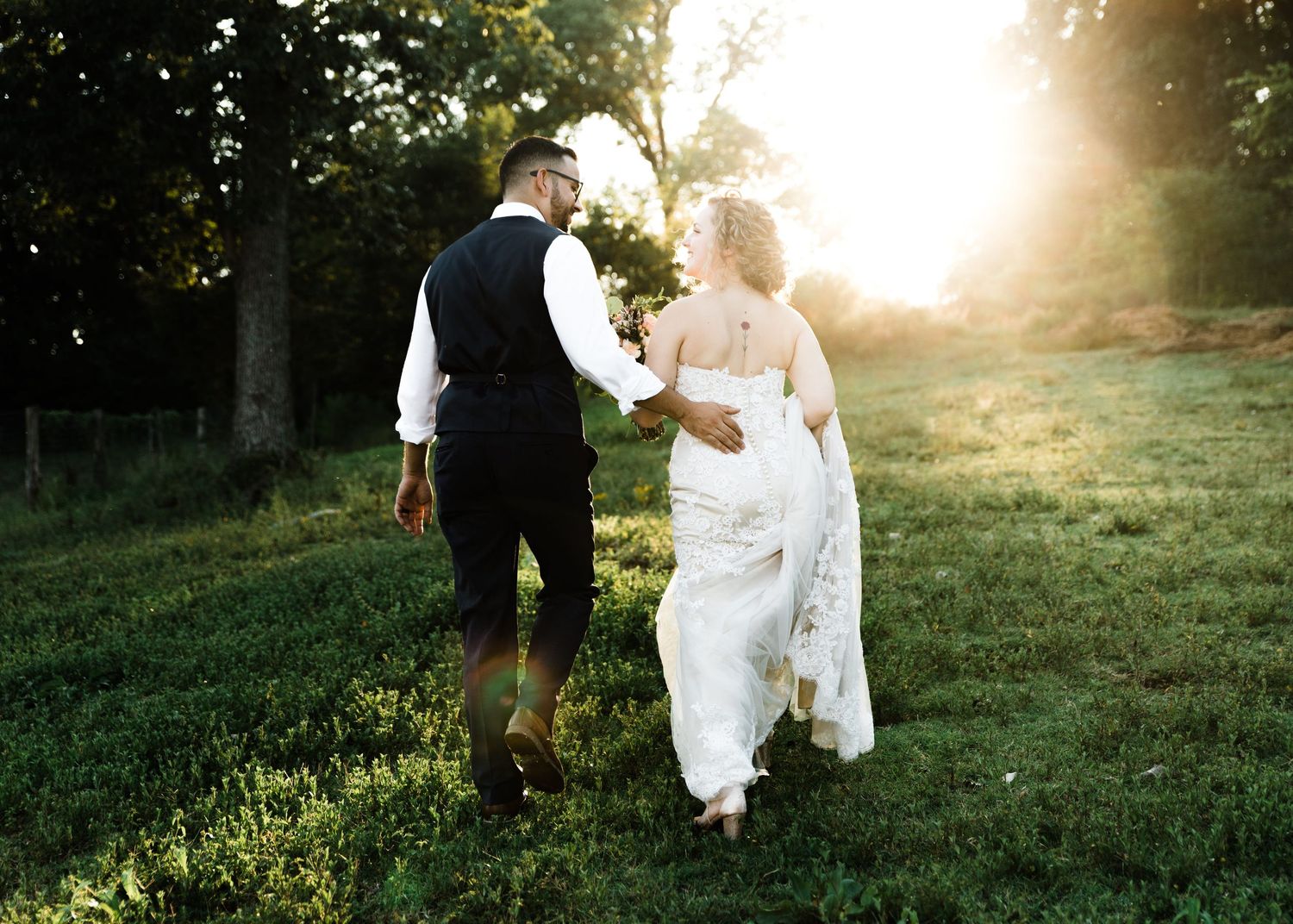 Bride and groom walking hand in hand through sunlit green meadow.