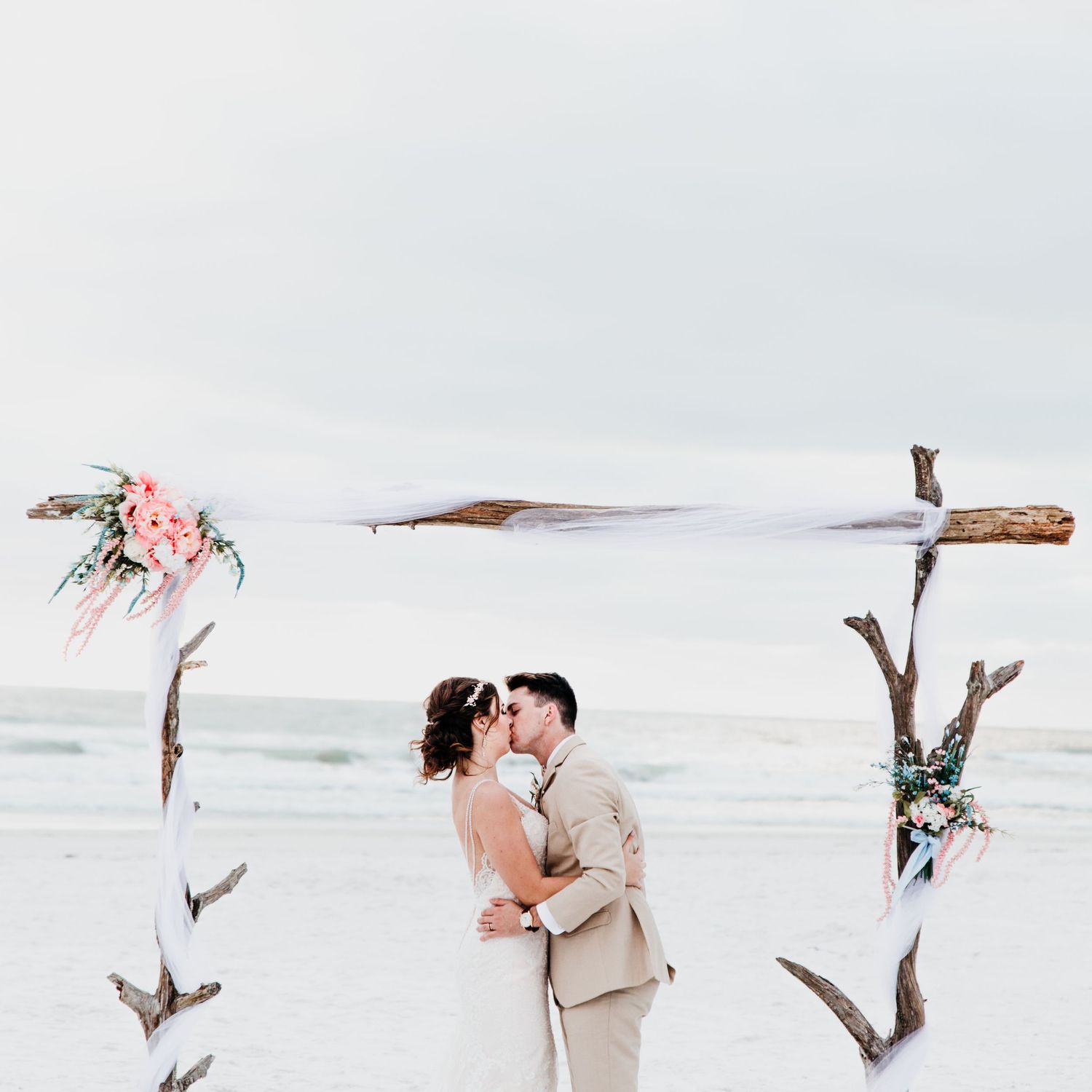 Couple kissing under driftwood arch decorated with flowers at a beach wedding ceremony.