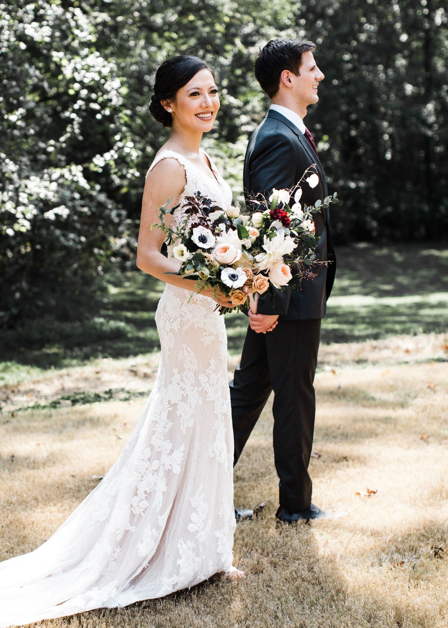Bride in lace gown holding anemone bouquet standing back-to-back with groom outdoors.