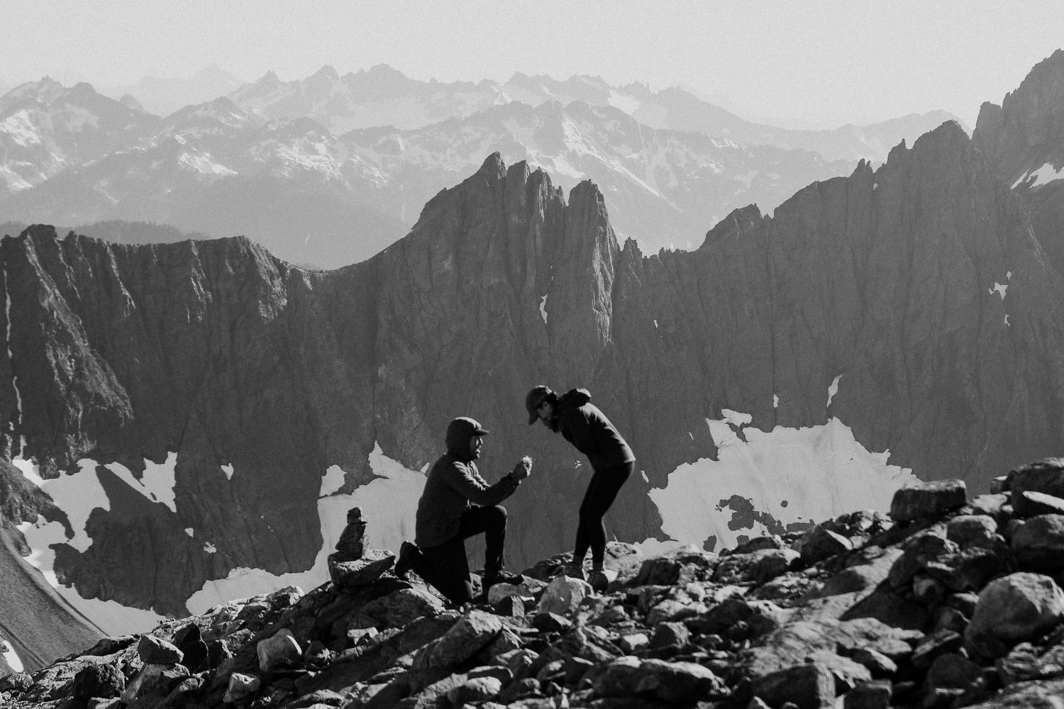 Black and white photo of a mountain proposal with a person kneeling before their partner on a rocky peak.