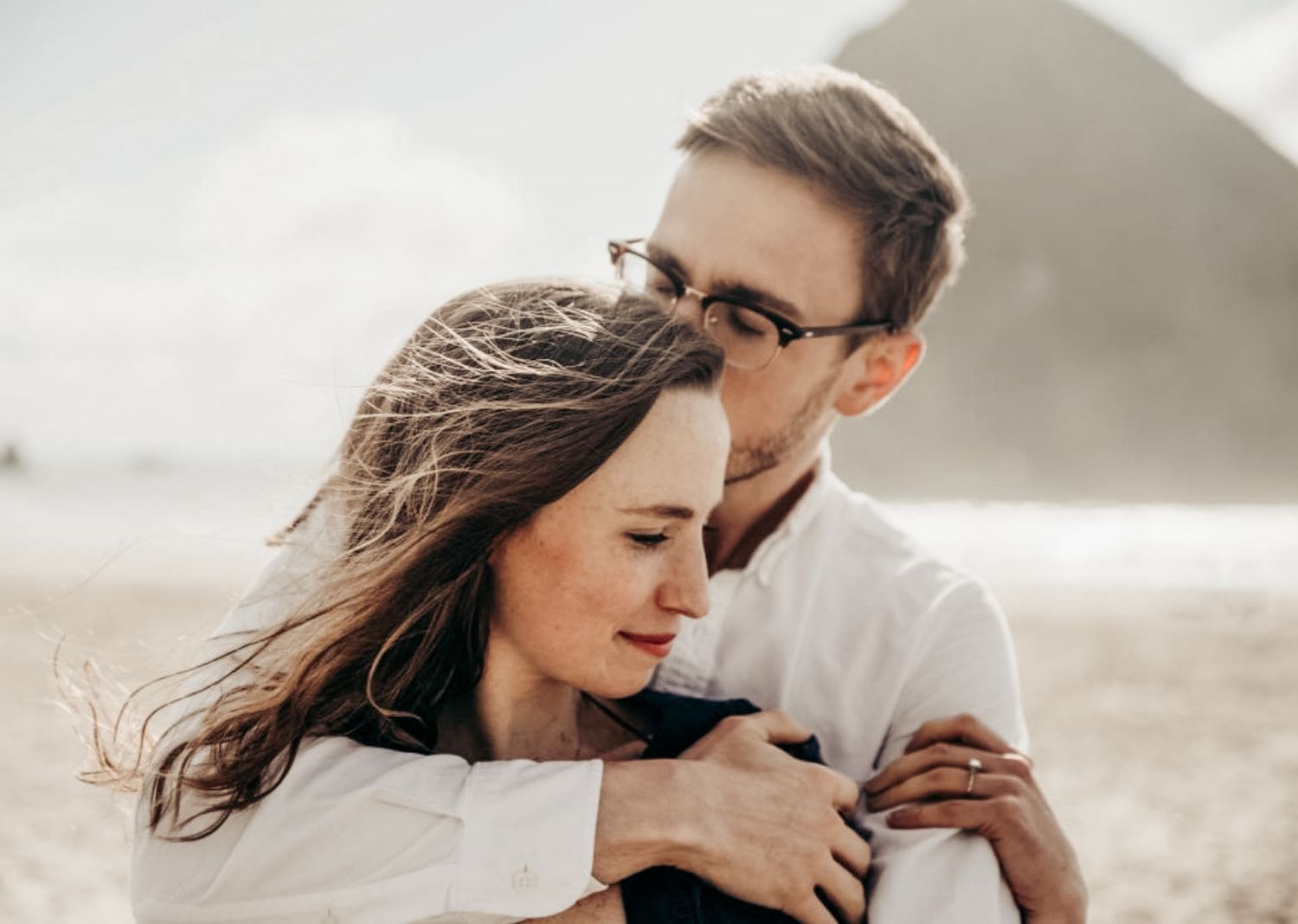 Tender moment of couple embracing on a windy beach, man in white shirt hugging woman from behind.