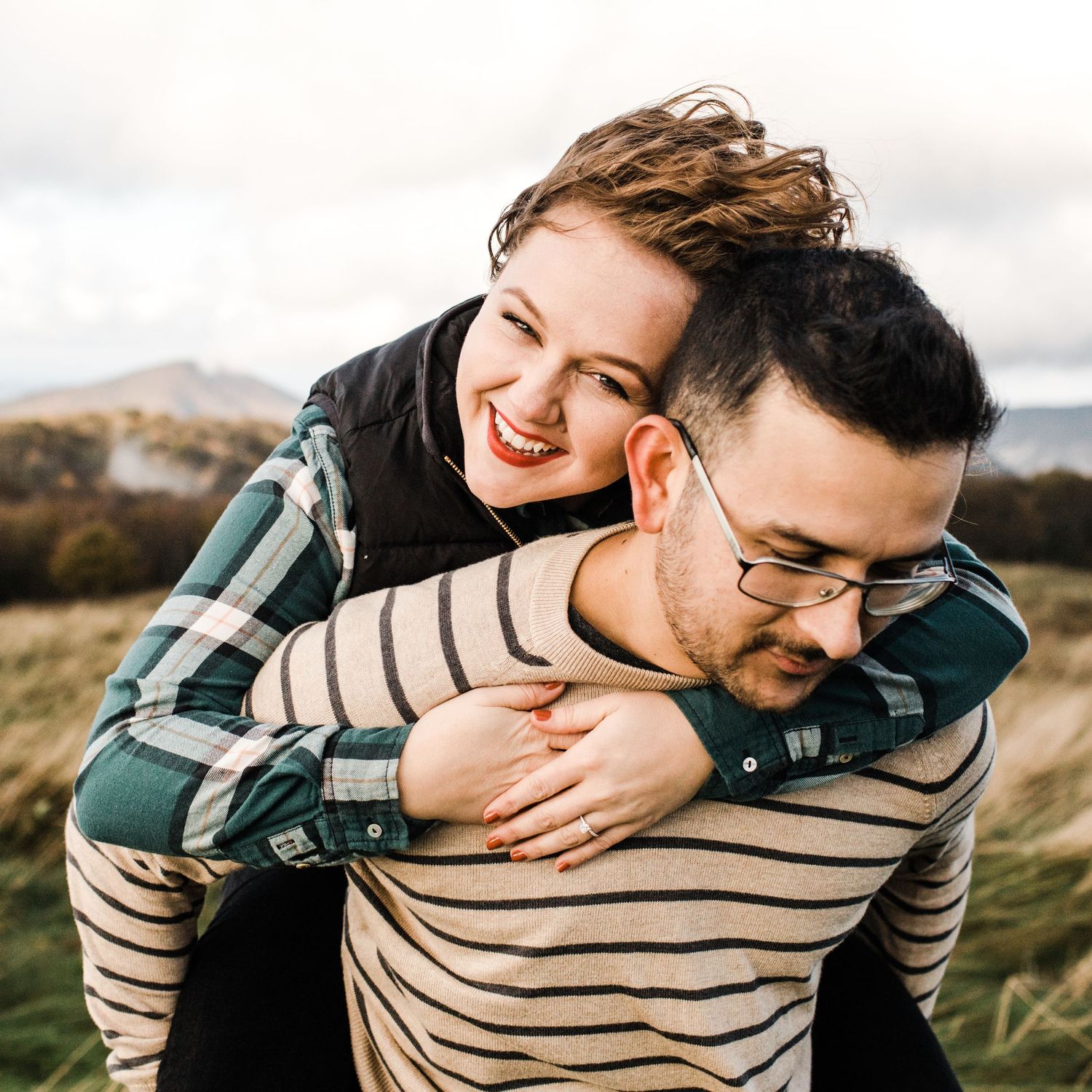 Joyful couple in casual outdoor engagement photo, woman piggyback riding man in field.