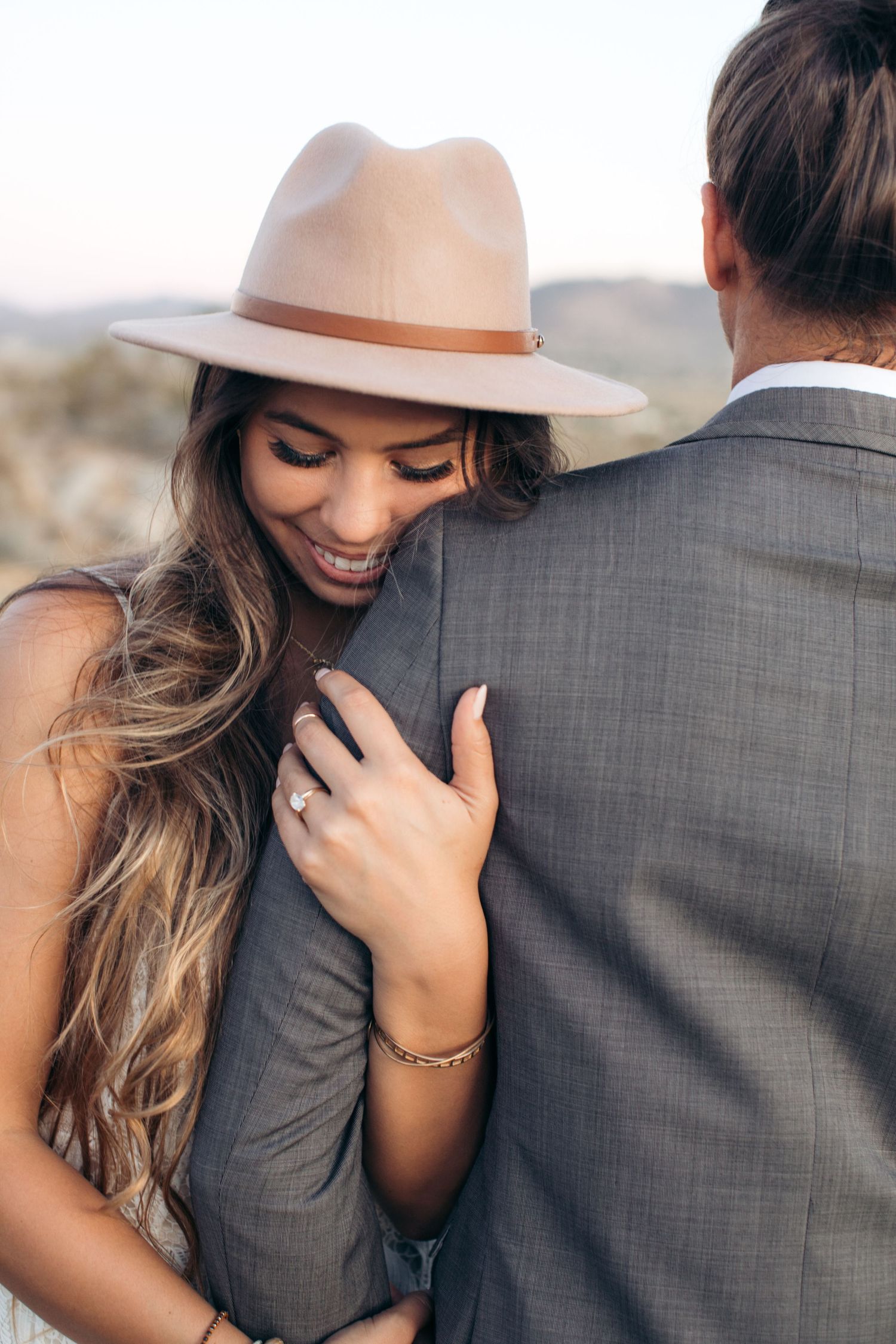 Smiling bride in a wide-brim hat embraces her groom in a gray suit during an outdoor engagement shoot.