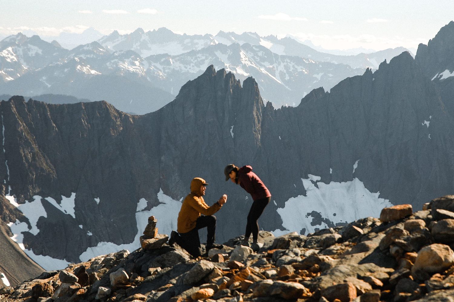 Man proposing to woman on a rocky mountain summit with stunning alpine peaks behind them.