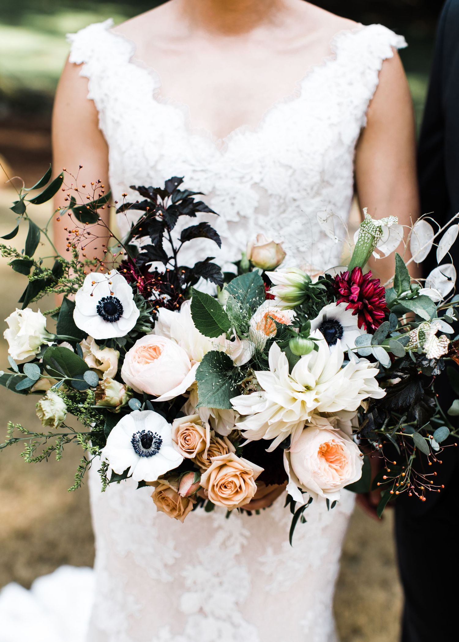 Bride holding a lush wedding bouquet with anemones, dahlias, roses, and eucalyptus in white, blush, and burgundy tones.