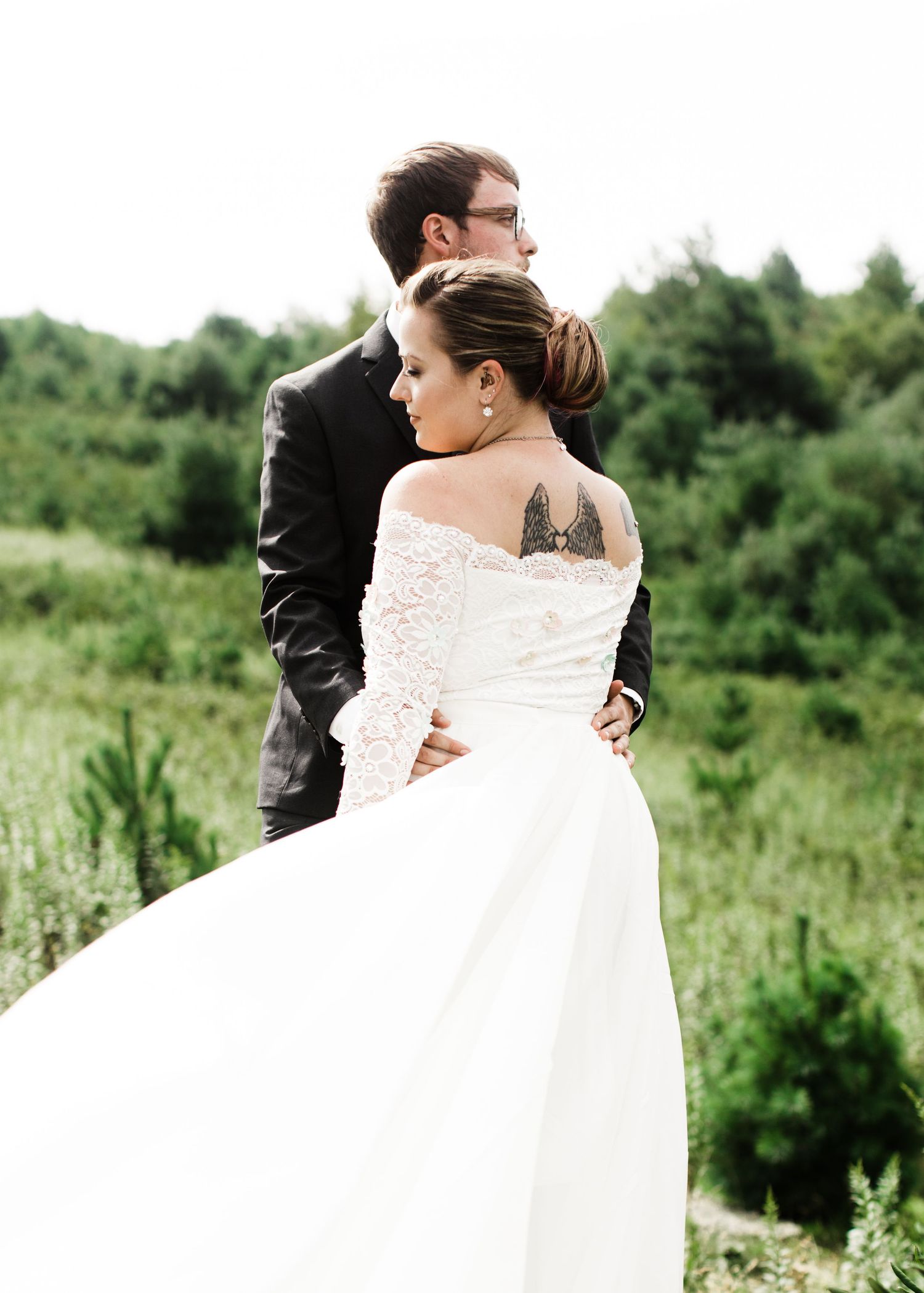 Bride in off-shoulder lace gown with back tattoo stands with groom in green field landscape.