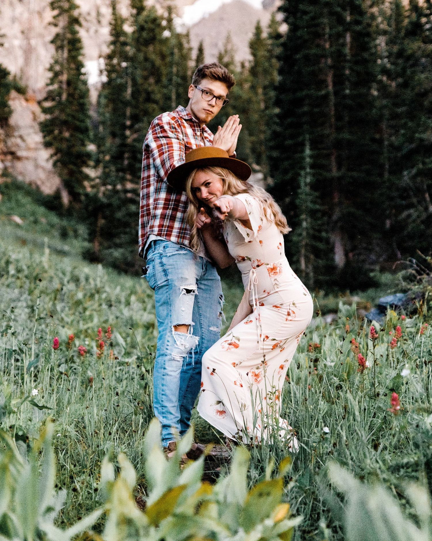 Couple posing playfully in a wildflower meadow with pine trees and mountains in the background.