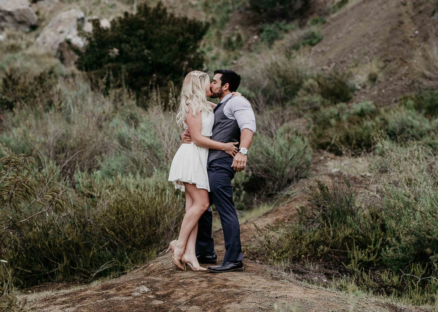 Couple sharing a romantic kiss on a rocky trail surrounded by lush green hillside.