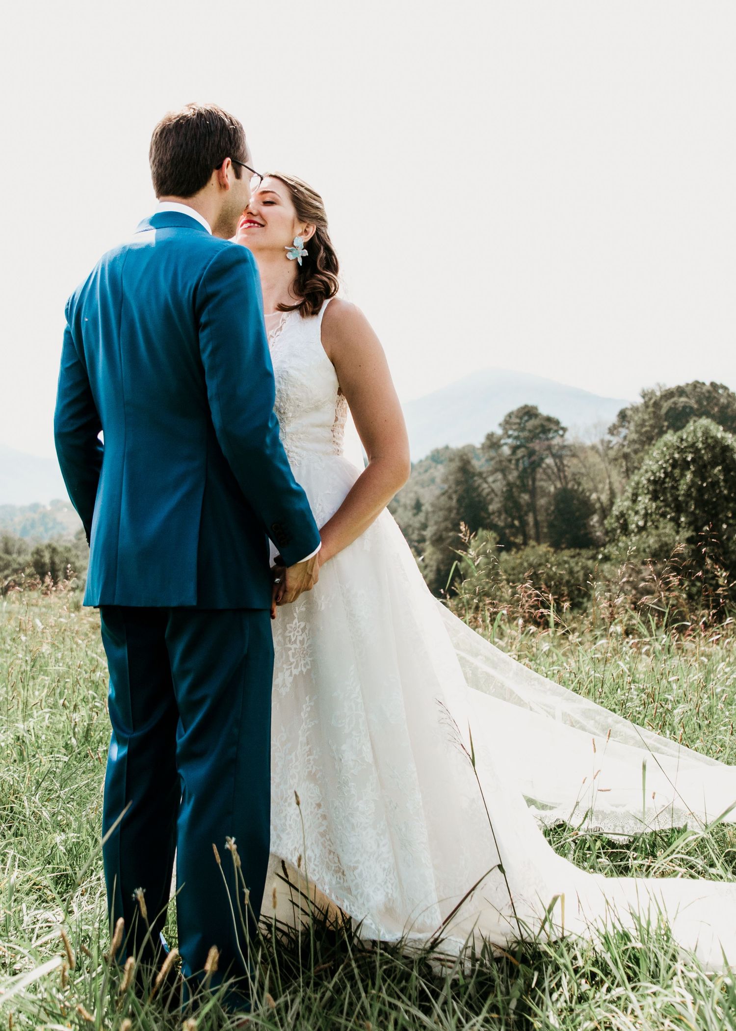 Bride in lace gown smiles at groom in blue suit as they hold hands in a scenic mountain meadow.