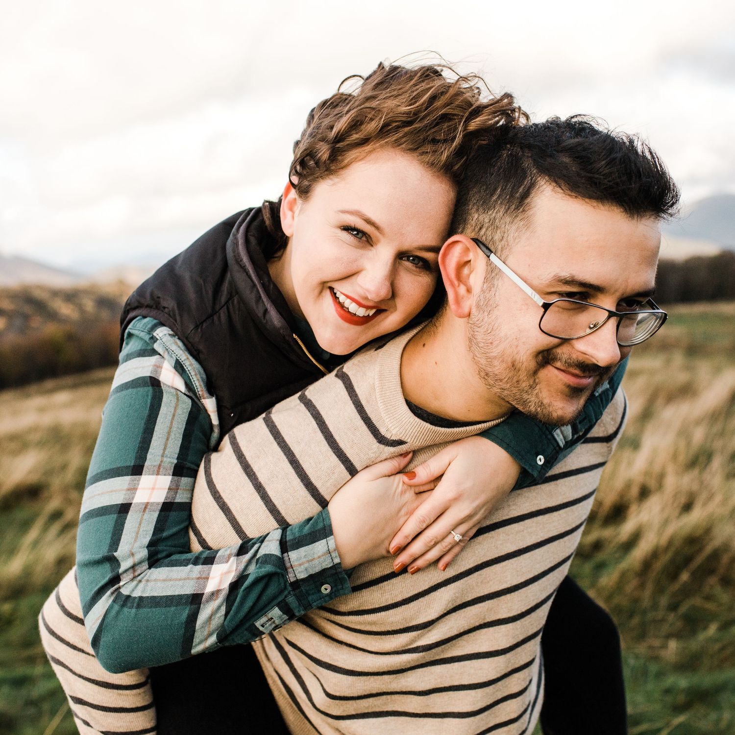 Happy couple laughing outdoors with woman giving piggyback ride in scenic countryside setting.