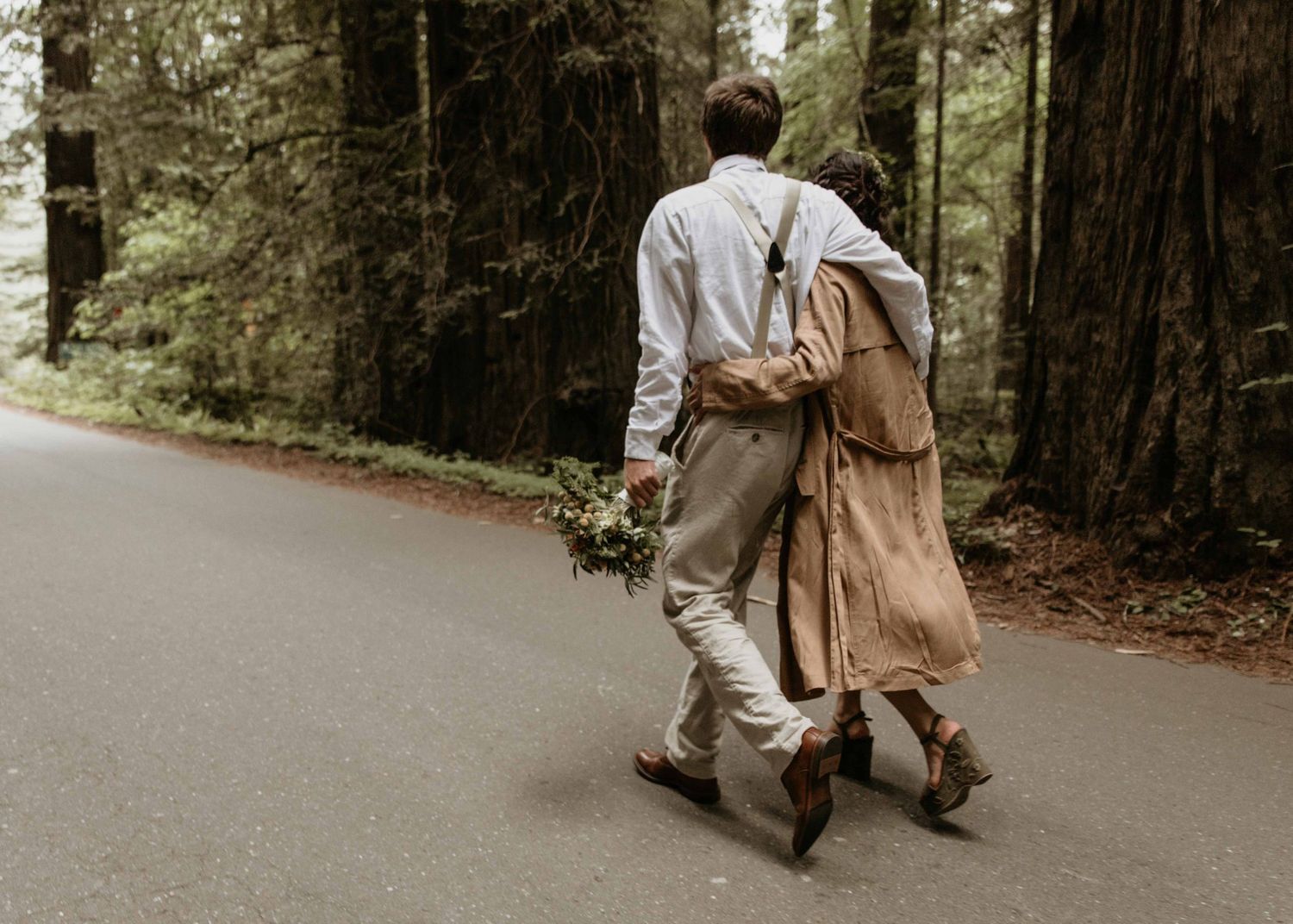 Newlywed couple walking hand in hand down a forest road, groom carrying a bouquet among tall redwoods.