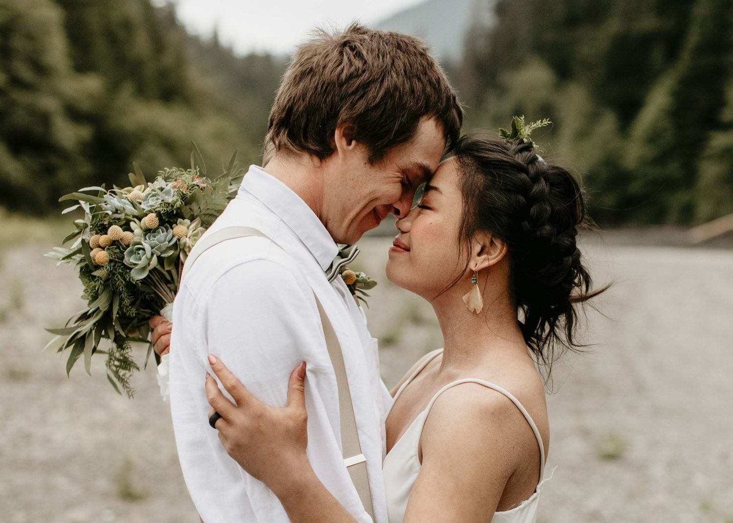 Romantic couple nearly kisses while groom holds a lush greenery bouquet near a mountain river.