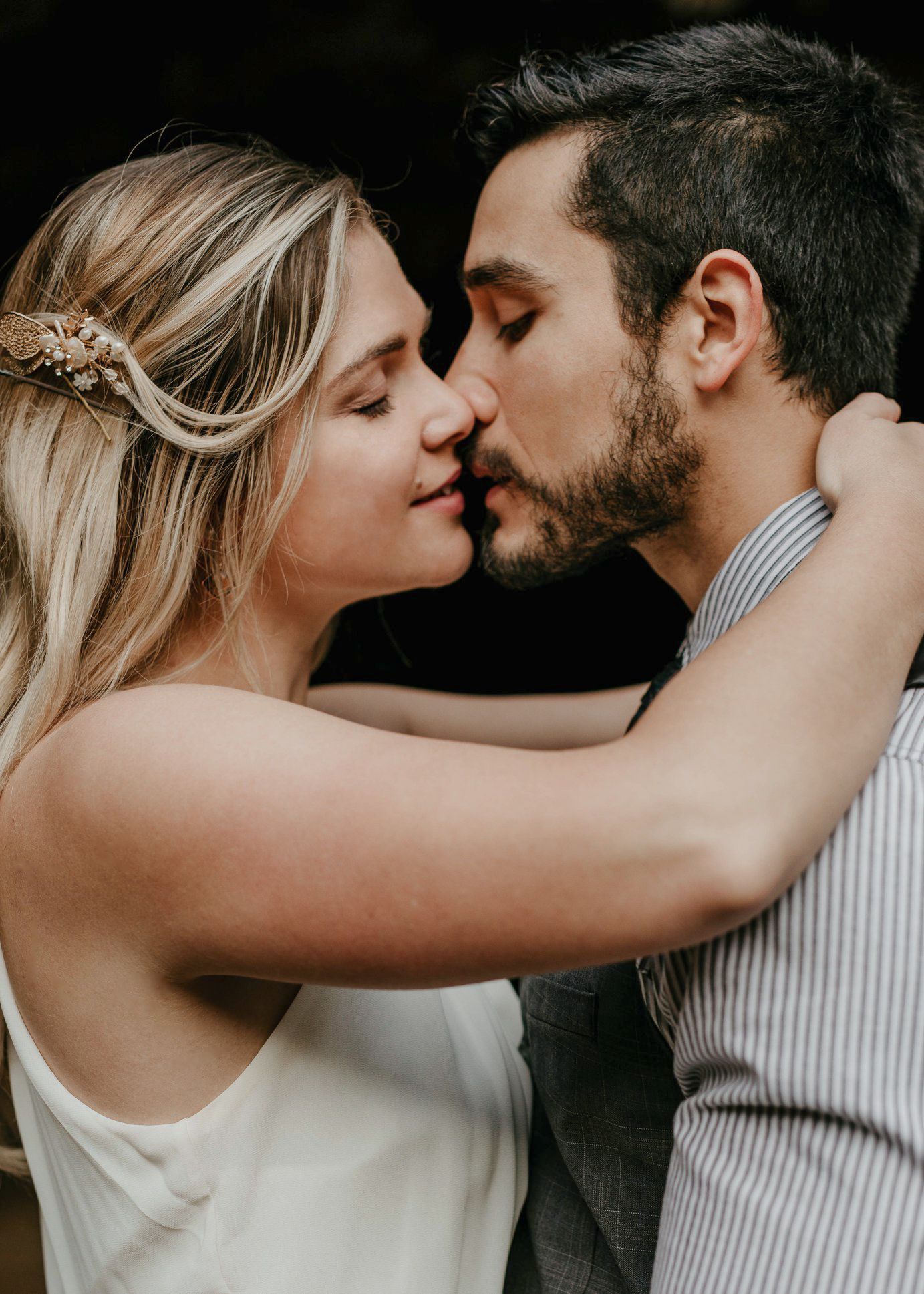 Romantic couple sharing a tender kiss, bride with floral hair accessory and groom in striped shirt.