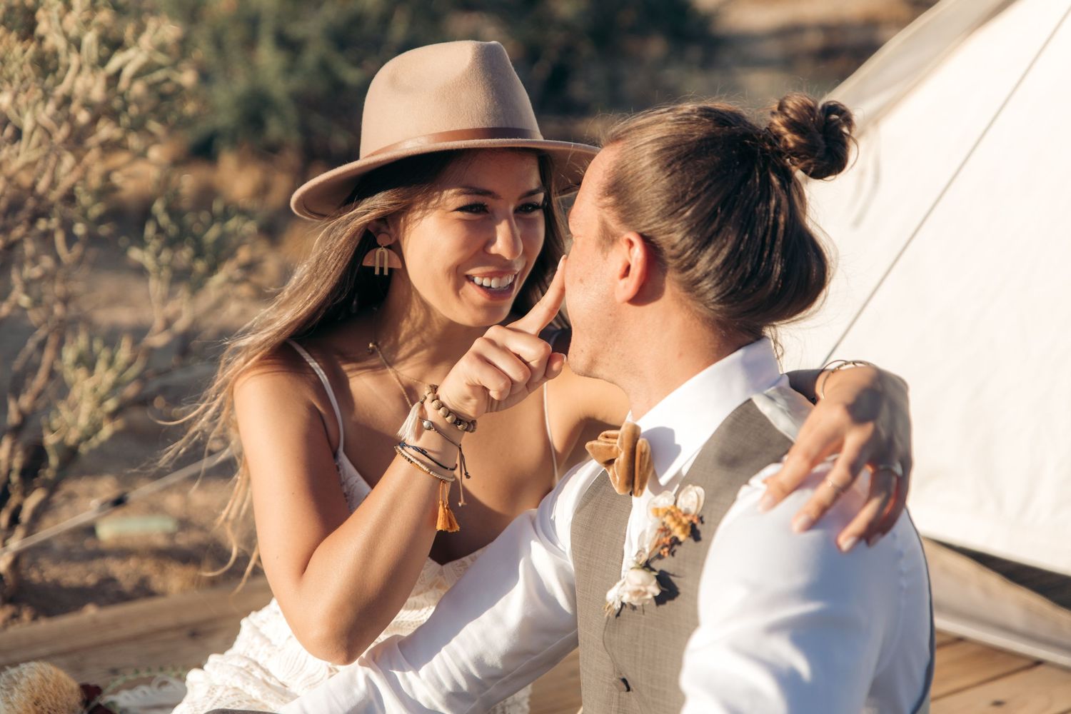 Smiling bride in boho hat laughs with groom near a glamping tent in a sunny desert setting.