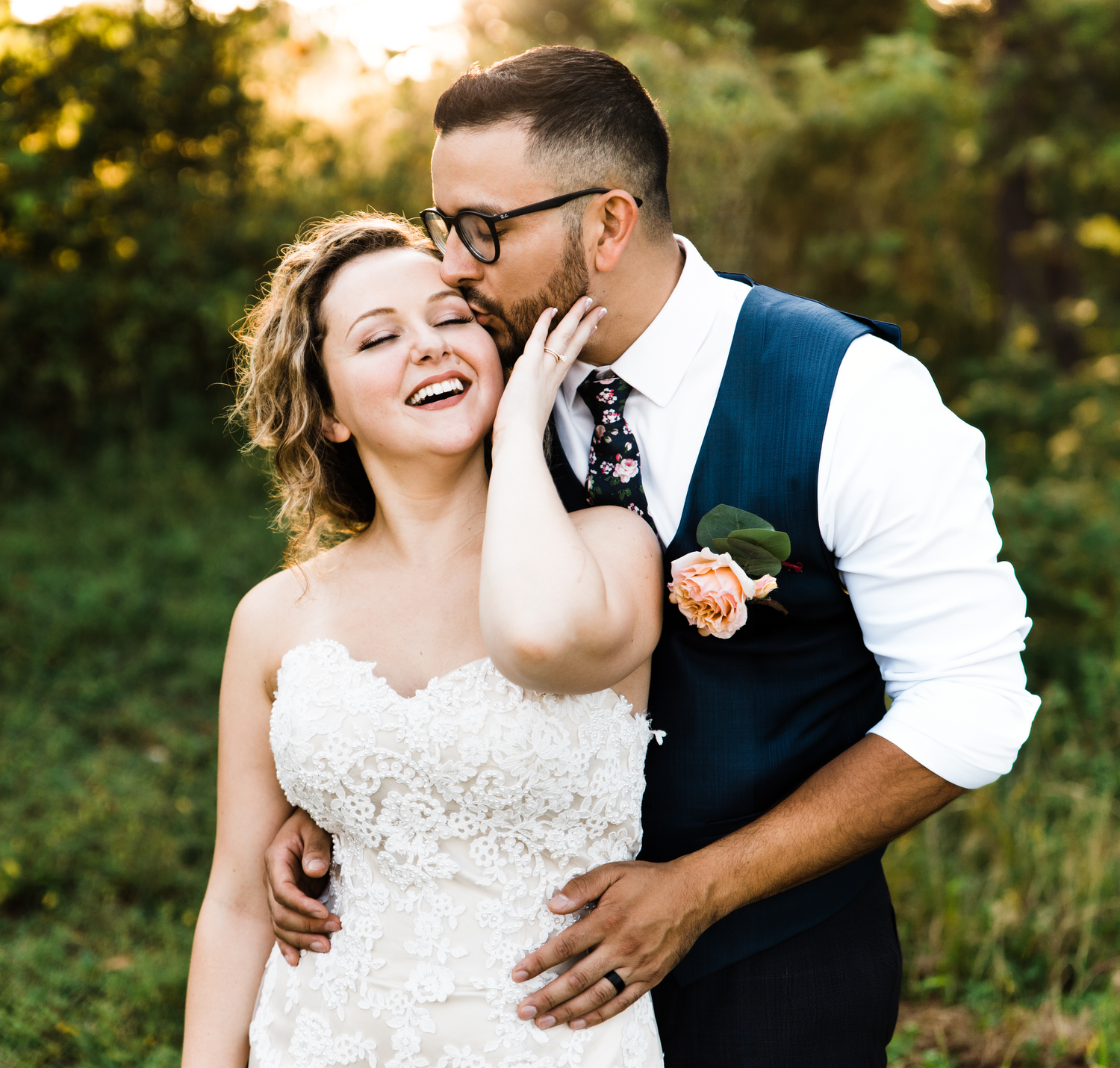 Laughing bride being kissed on the cheek by groom in a navy vest outdoors in golden light.
