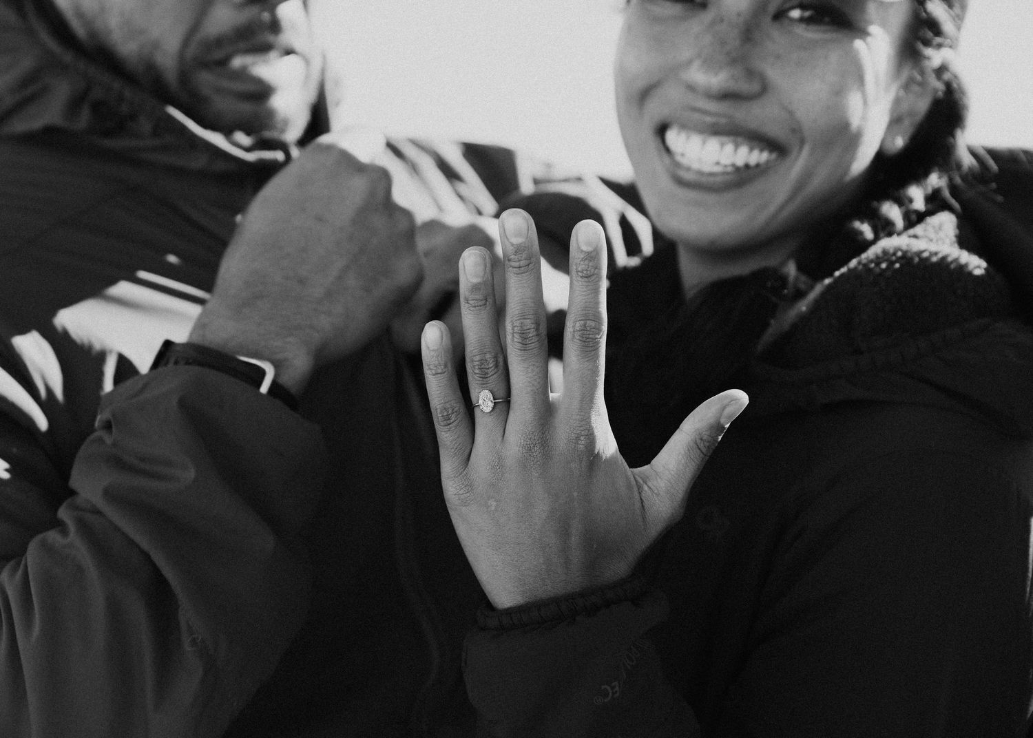 Black and white photo of joyful woman displaying diamond engagement ring with partner.