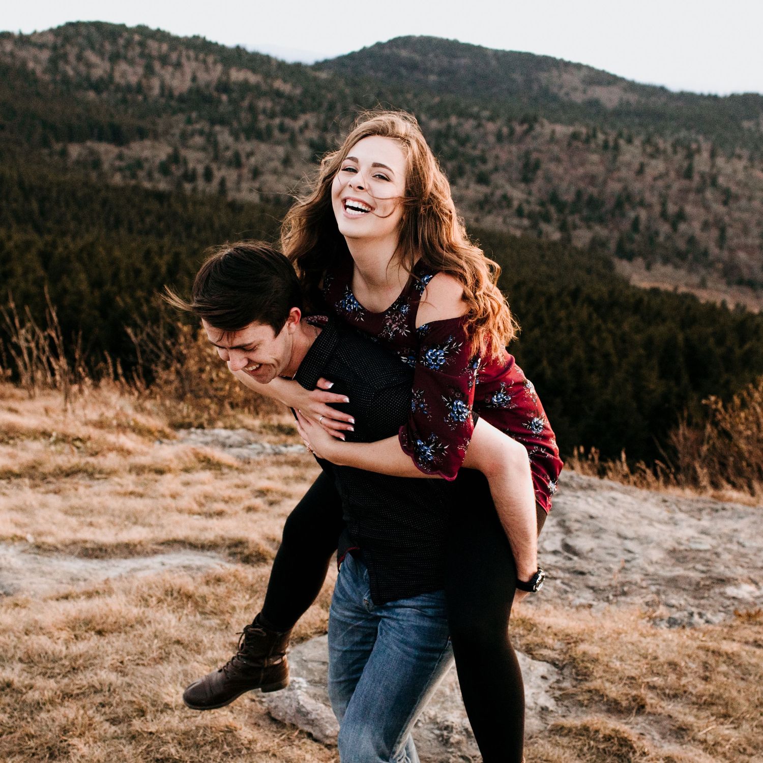 Joyful couple laughing on mountain hillside with man giving woman a piggyback ride.