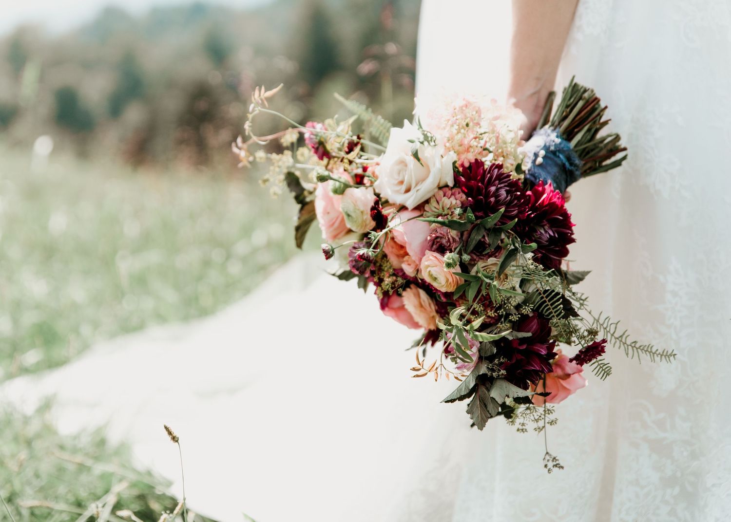 Bride holds a lush bridal bouquet of burgundy, pink, and white blooms with greenery in a field.