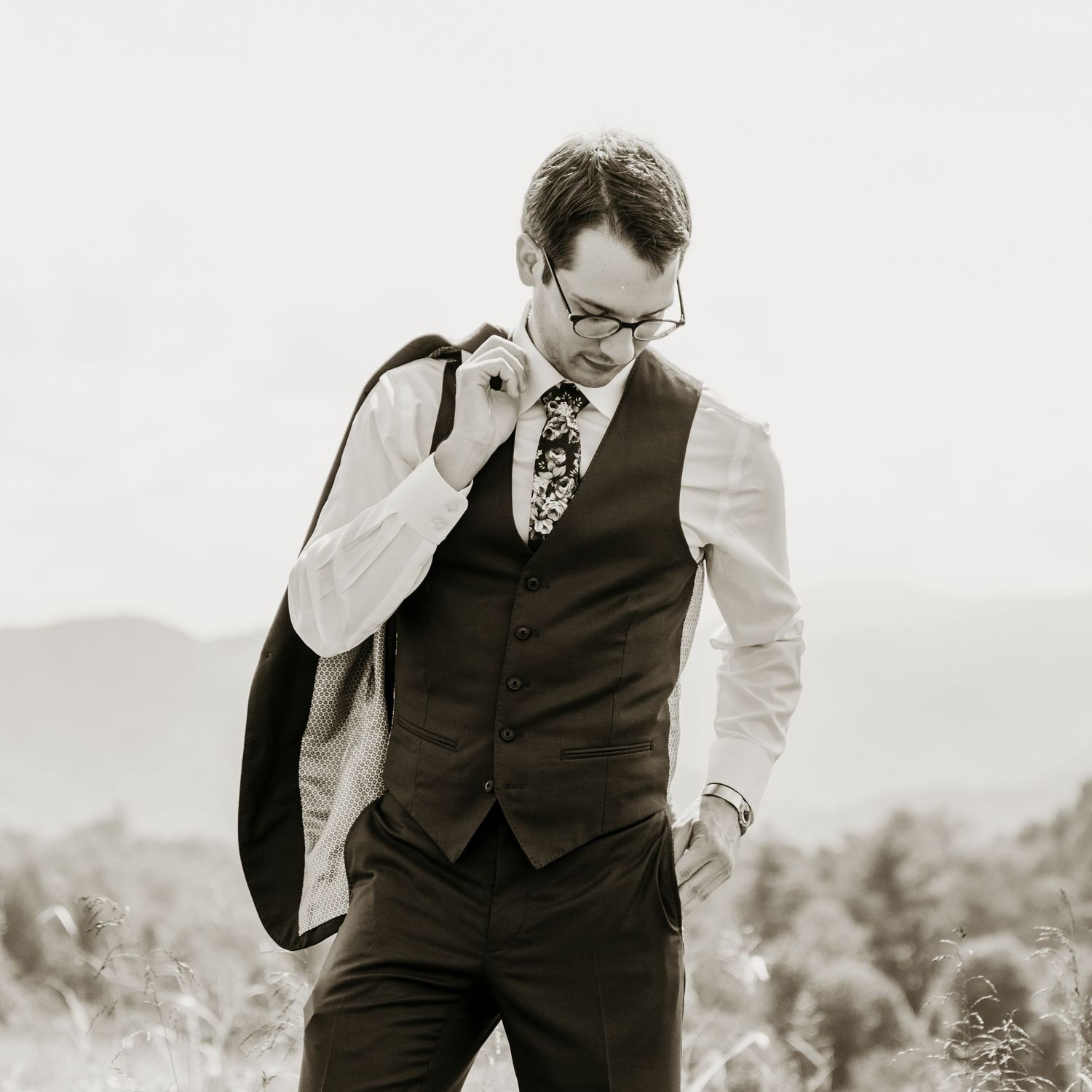 Black and white portrait of a stylish groom in a vest and floral tie holding his jacket over his shoulder.