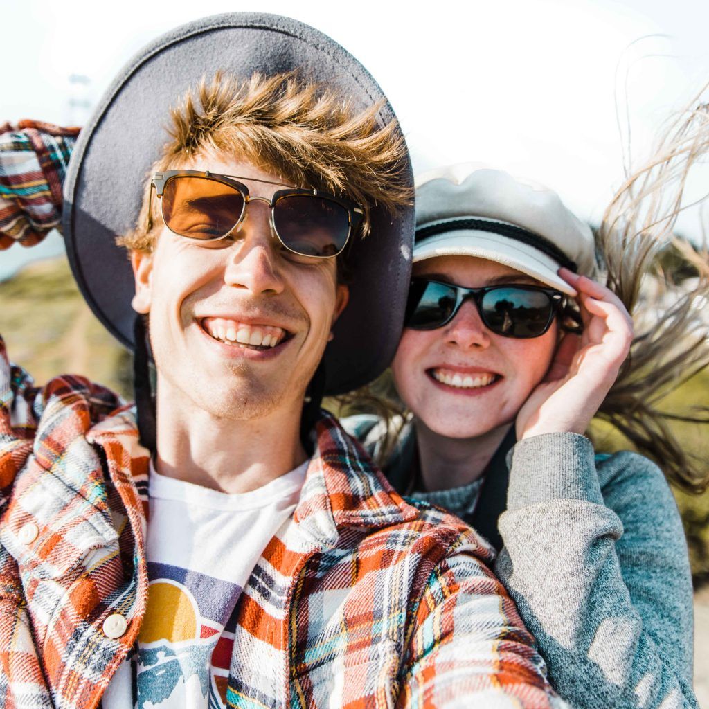 Happy couple wearing sunglasses and hats taking a fun selfie outdoors on a windy day.