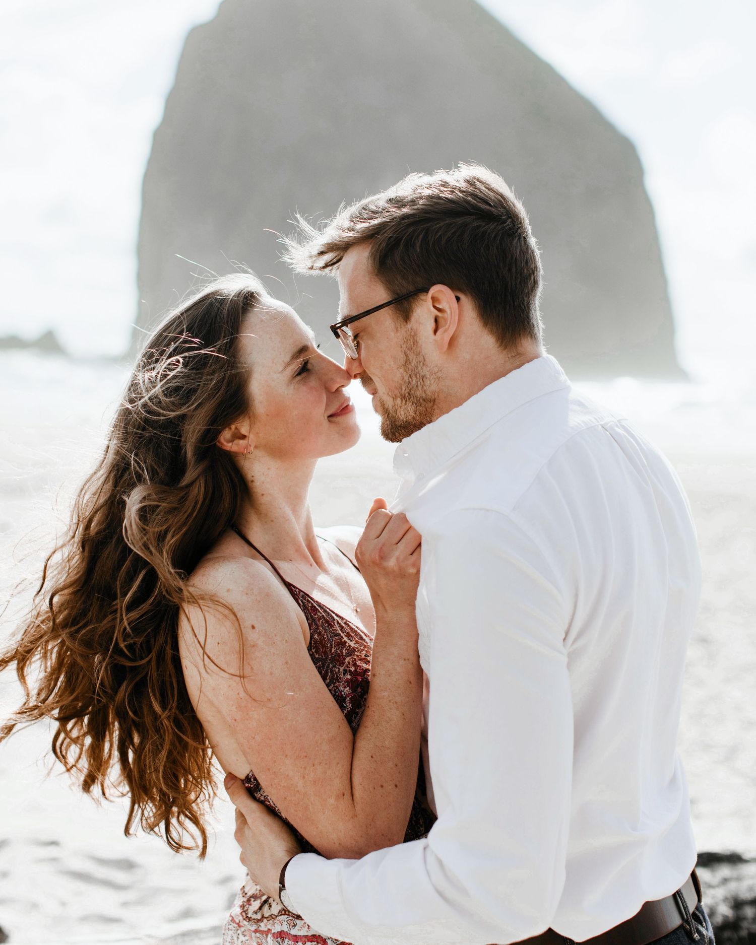 Romantic couple nearly kisses on a windswept beach with iconic Haystack Rock in the background.