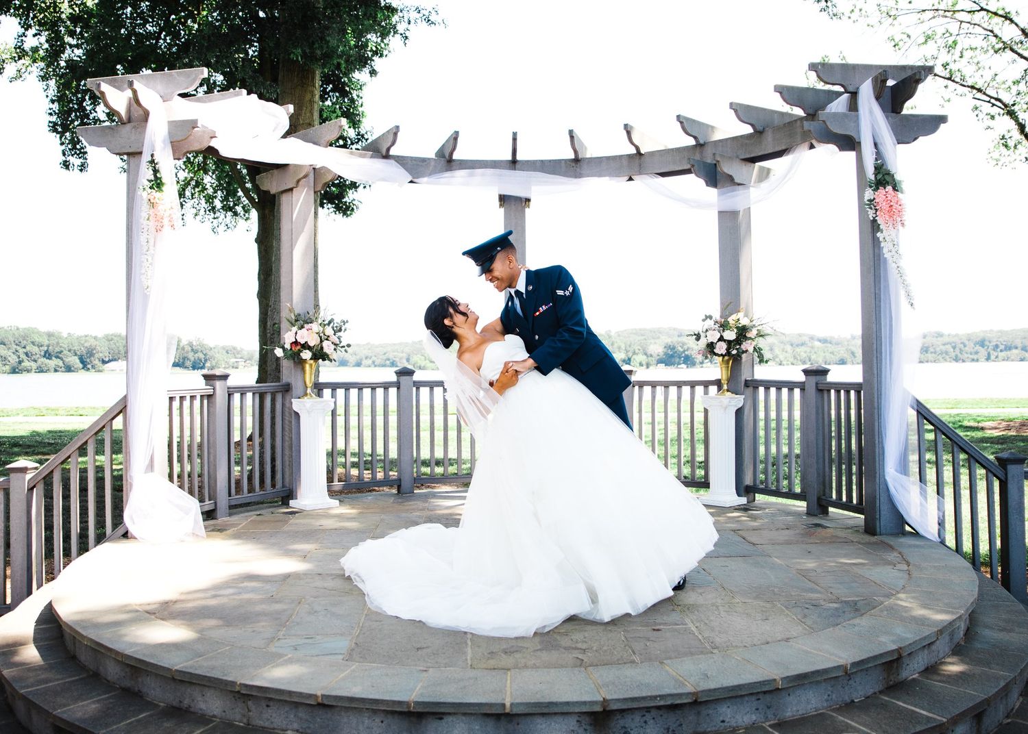 Groom dips bride romantically under elegant pergola with scenic waterfront backdrop.