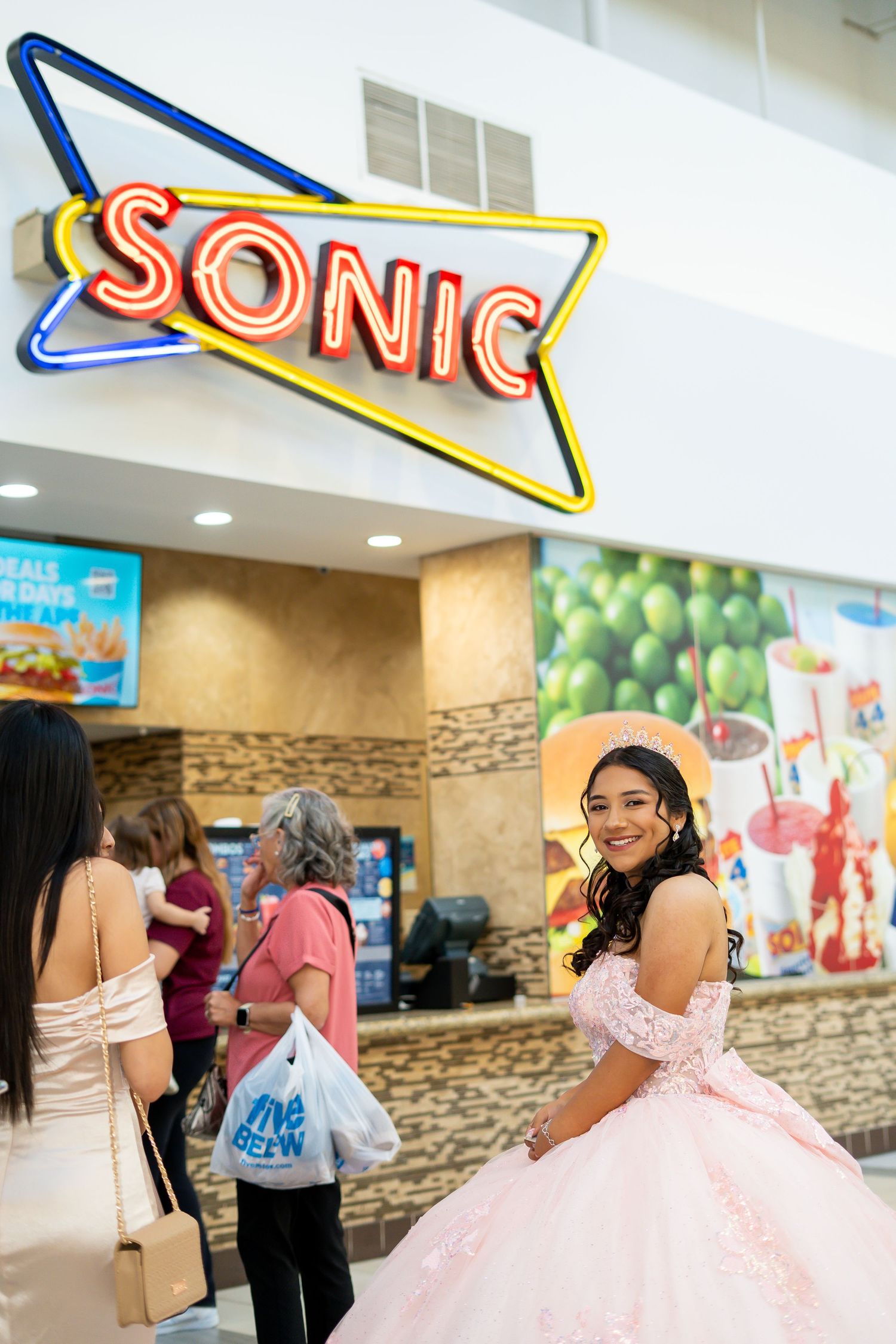 Quinceañera in pink ball gown smiling in front of a Sonic restaurant neon sign inside a mall.