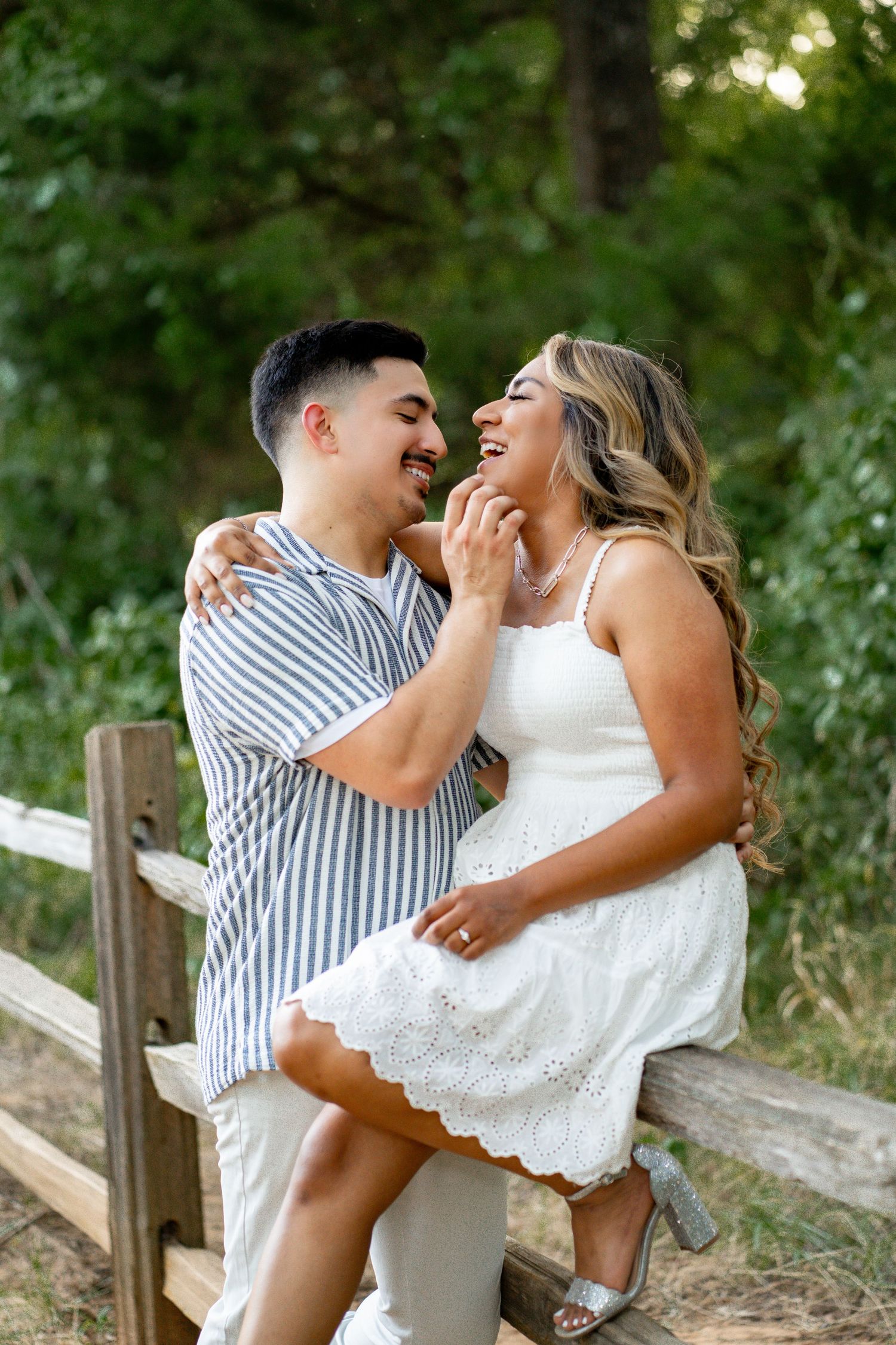 Laughing couple sitting on a wooden fence together in a lush green outdoor setting.