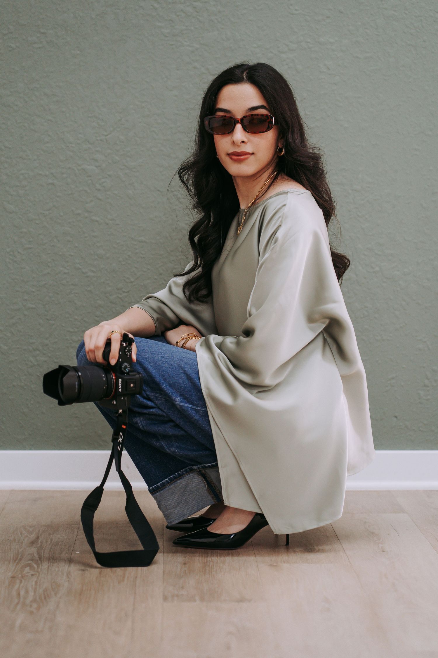 Woman in sage tunic and jeans sits against green wall holding a DSLR camera, wearing brown sunglasses.