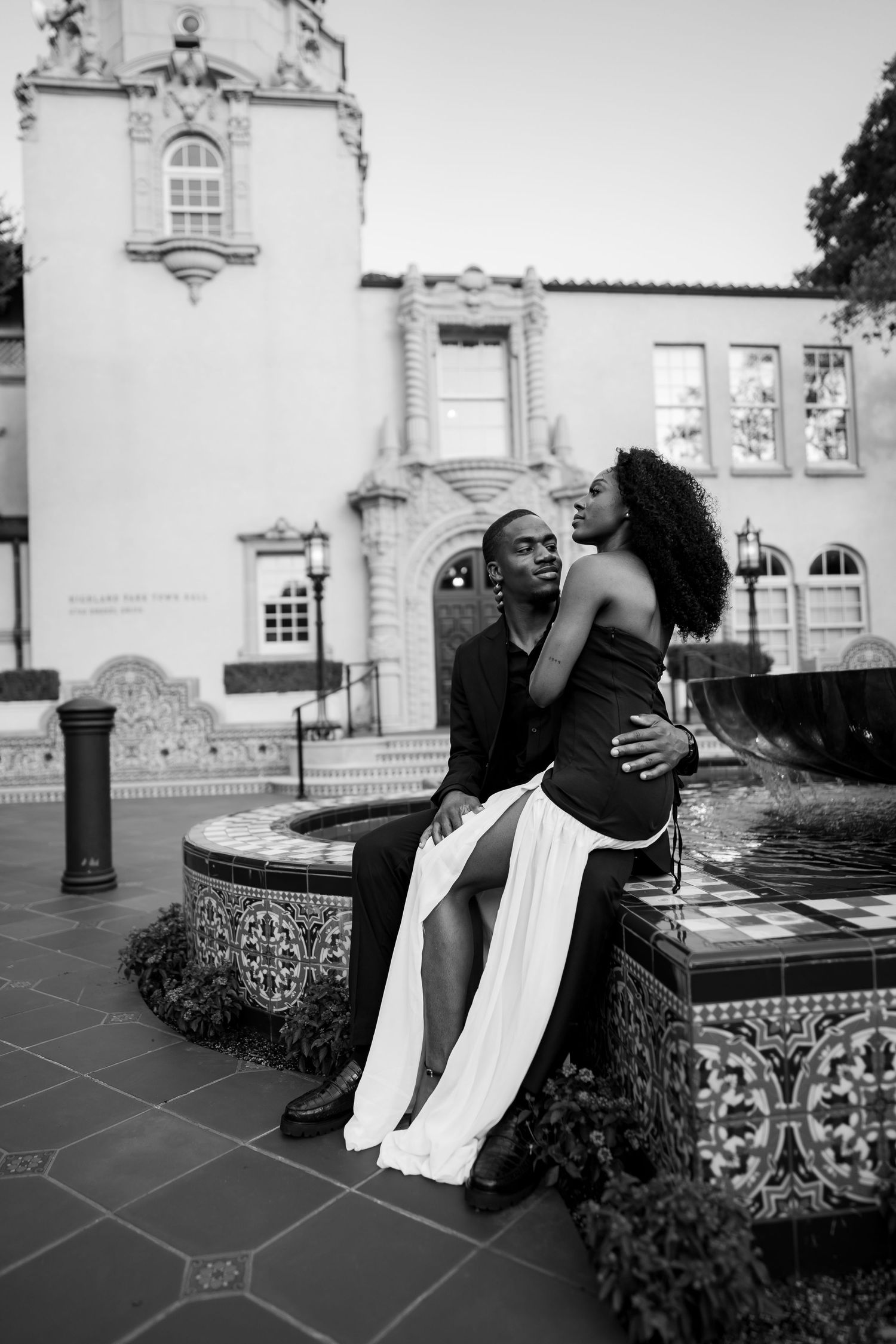 Black and white romantic photo of a couple embracing by a tiled fountain outside a Spanish-style building.