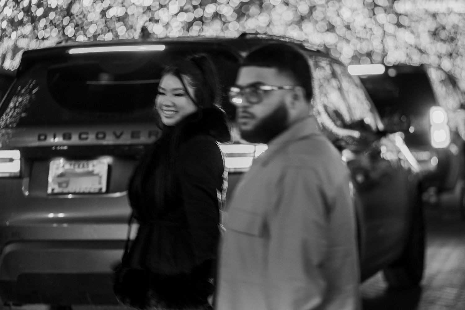 Couple walking past a Land Rover Discovery with festive lights in the background, black and white photo.