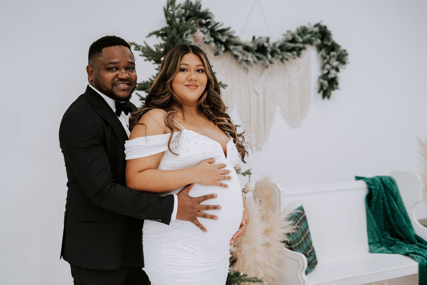 Couple maternity photo with man in tuxedo embracing pregnant woman in white gown before macrame backdrop.