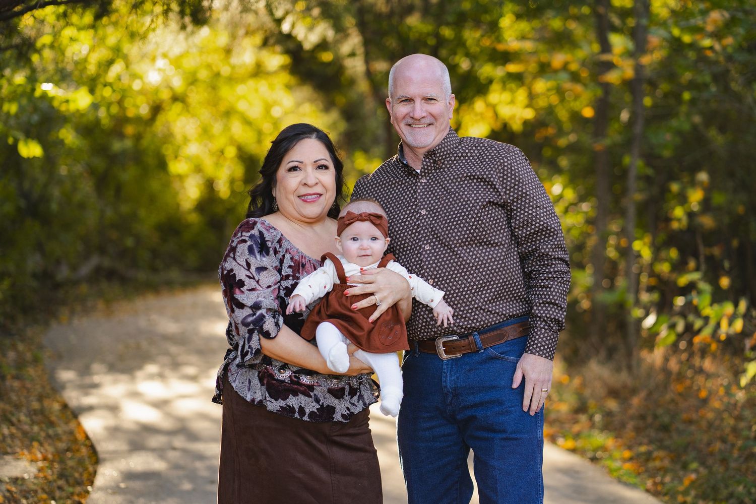 Grandparents proudly holding a baby in a rust-colored outfit on a scenic fall nature path.